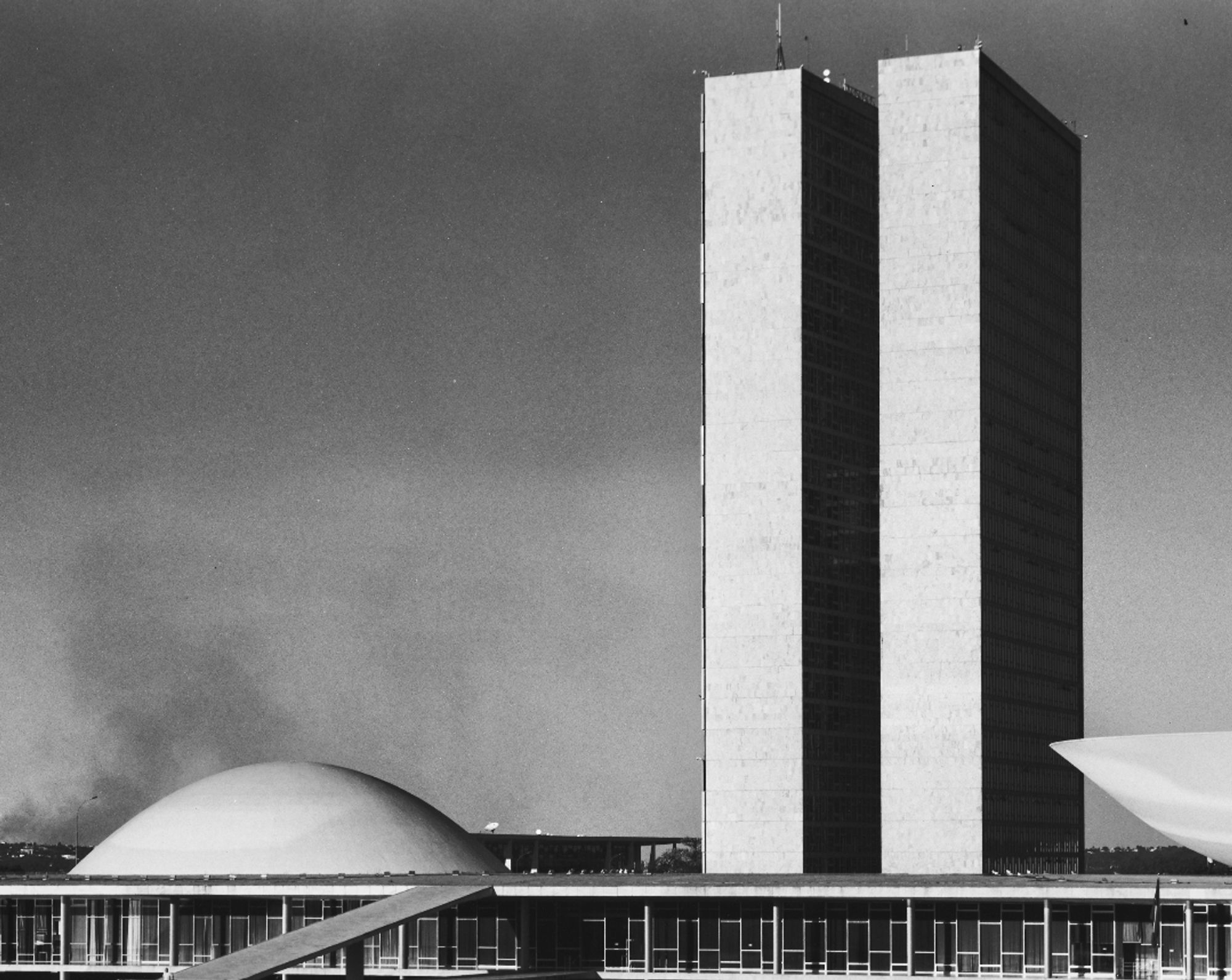 Prédio do Congresso Nacional com foco na parte do Senado Federal e, ao fundo, fumaça subindo e acumulando no céu, como se saísse da cúpula do Senado.