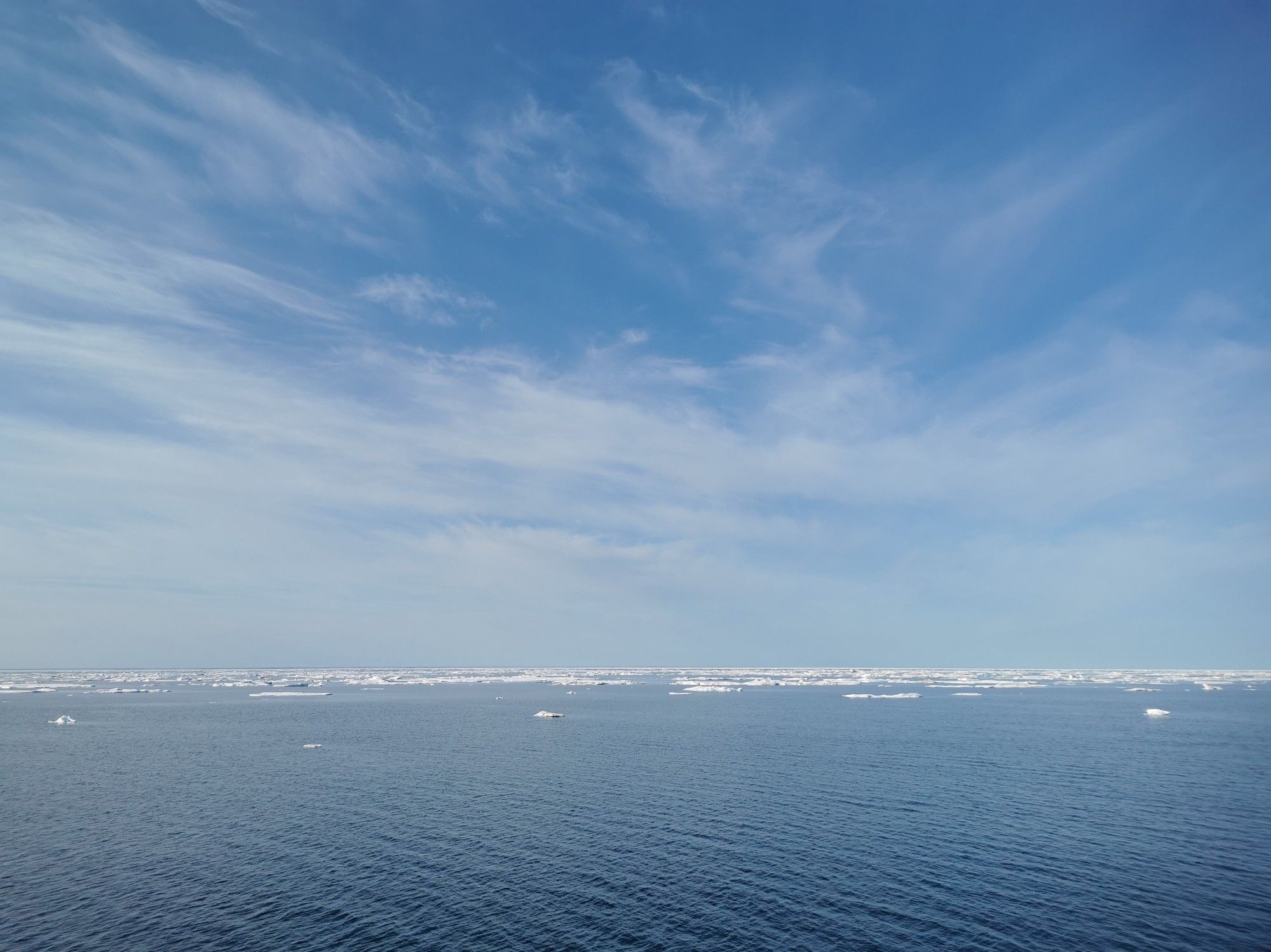 A sea view with blue skies, fluffy white clouds, calm water and scattered pack ice towards the horizon.