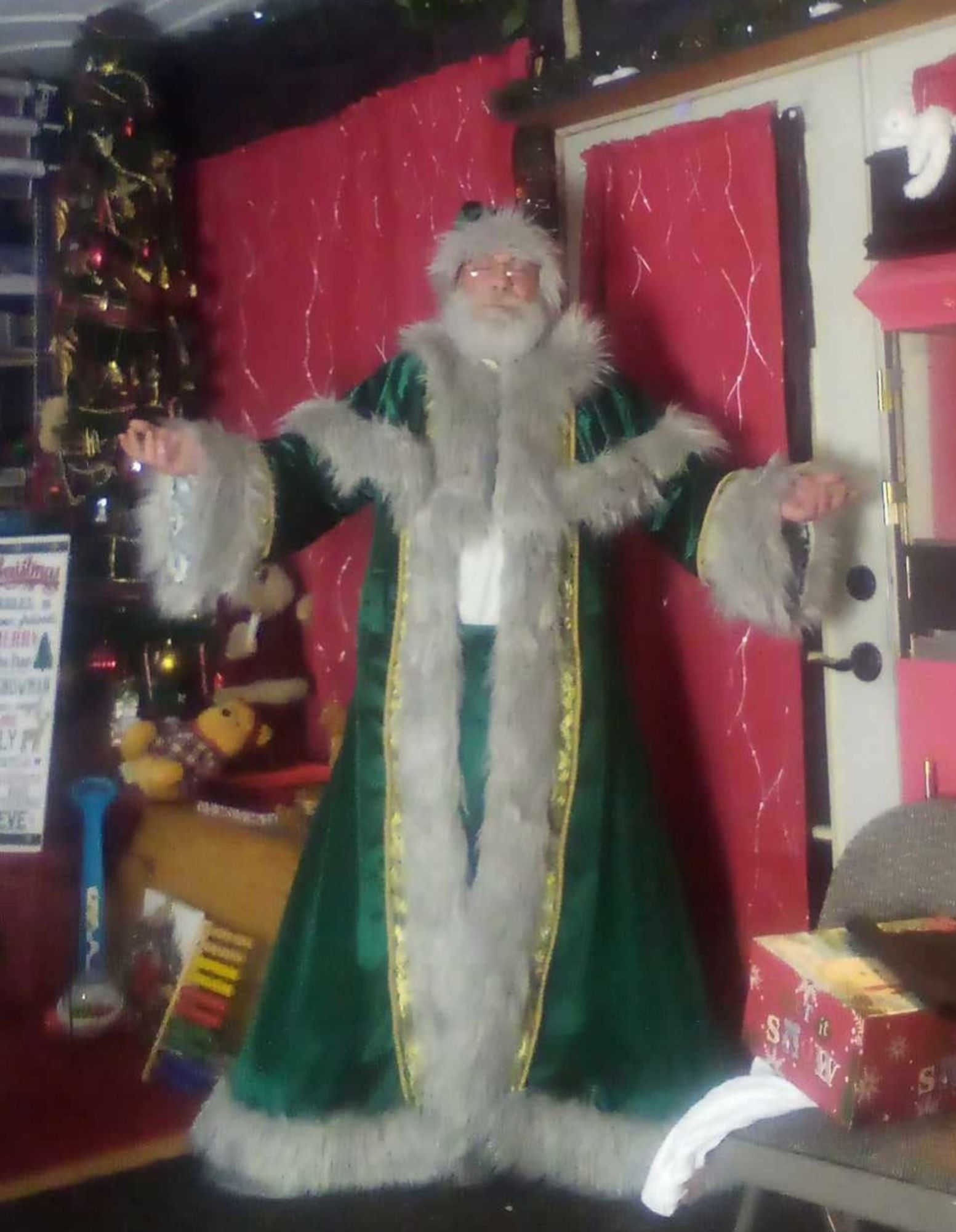 A man in a full-length green velvet robe with green capelet and hat, with silver fur, in front of a Christmas inspired background.