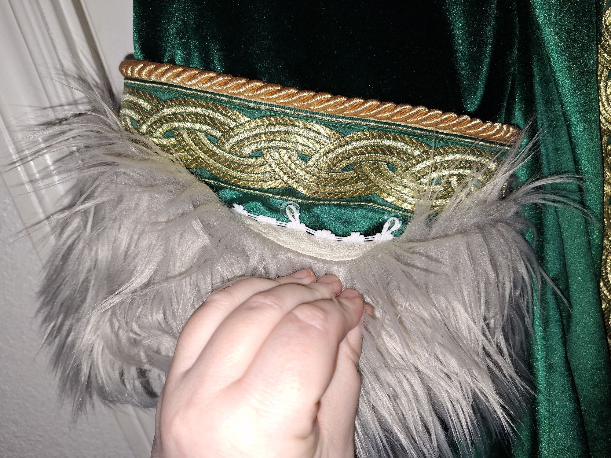 The end of a green velvet robe with gold trim, with the fur trim pulled back to reveal a button and loop connection system for easy removal.