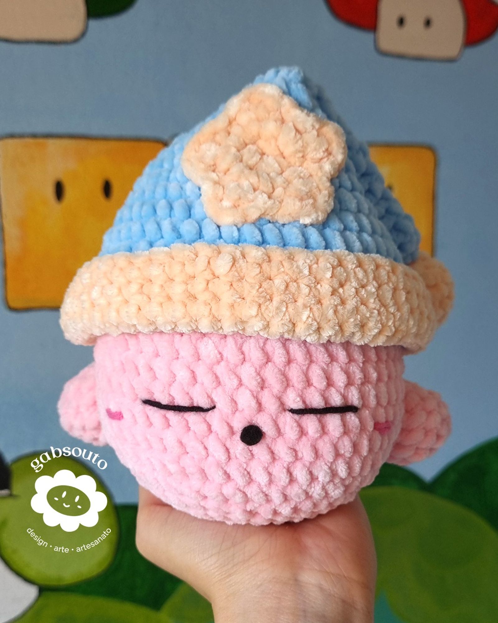 Uma mão segura um amigurumi do personagem Kirby, com uma expressão sonolenta, utilizando um gorro de pelúcia azul, com uma estrela amarela no meio e bola na amarela na ponta. Ao fundo, uma parece com desenhos do jogo Mario.