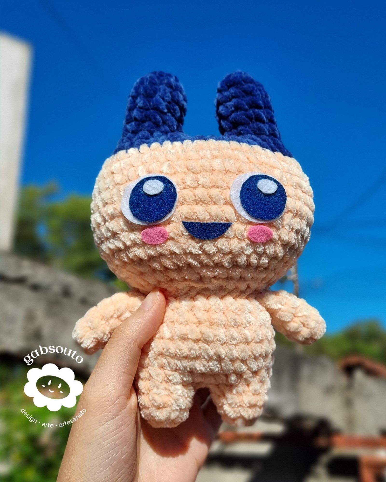 Foto de um amigurumi do personagem Mametchi do jogo Tamagotchi. Mão segurando o amigurumi, num fundo com um muro e o céu.