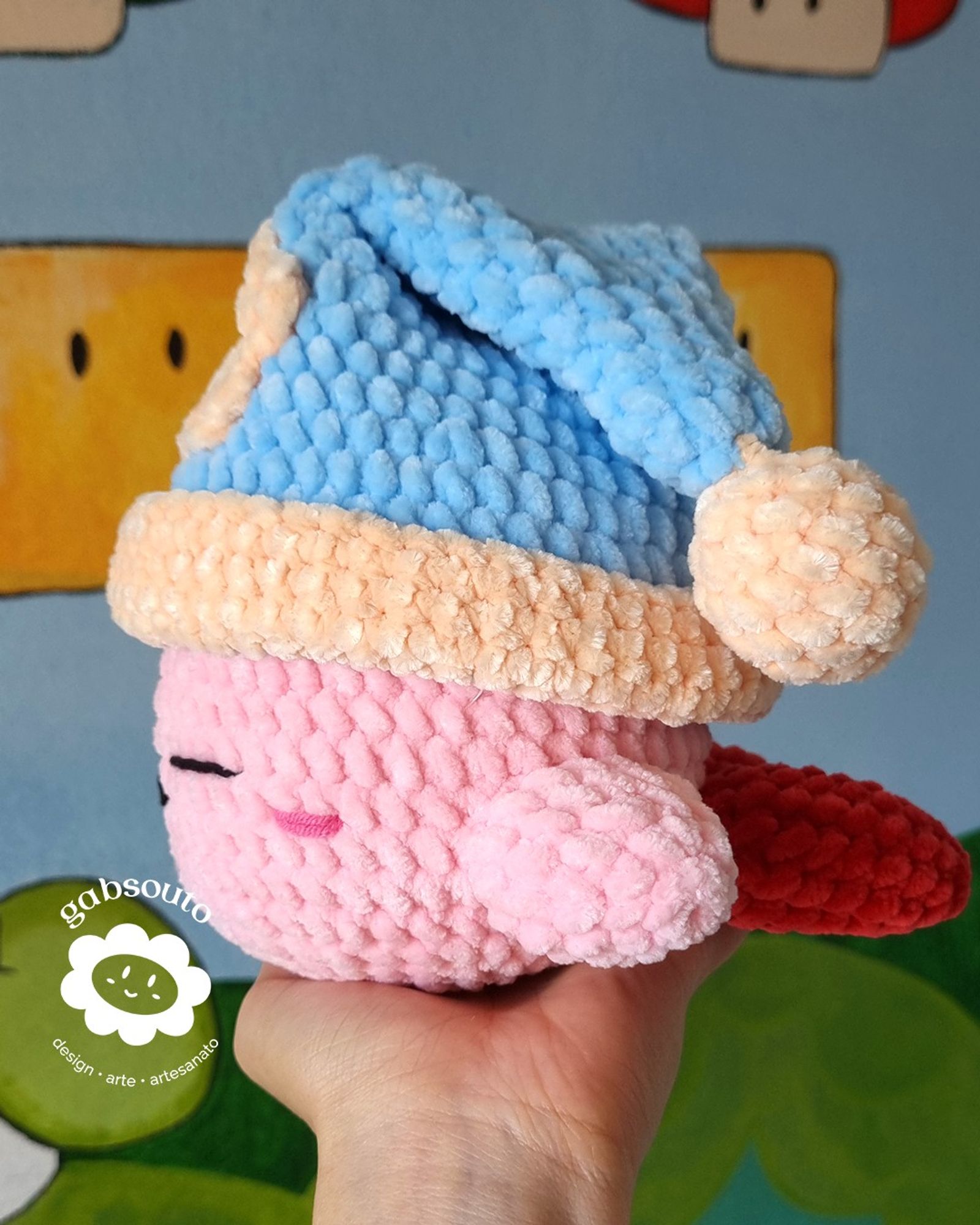 Uma mão segura um amigurumi em perfil, do personagem Kirby utilizando um gorro de pelúcia azul, com uma bola na amarela na ponta. Ao fundo, uma parece com desenhos do jogo Mario.