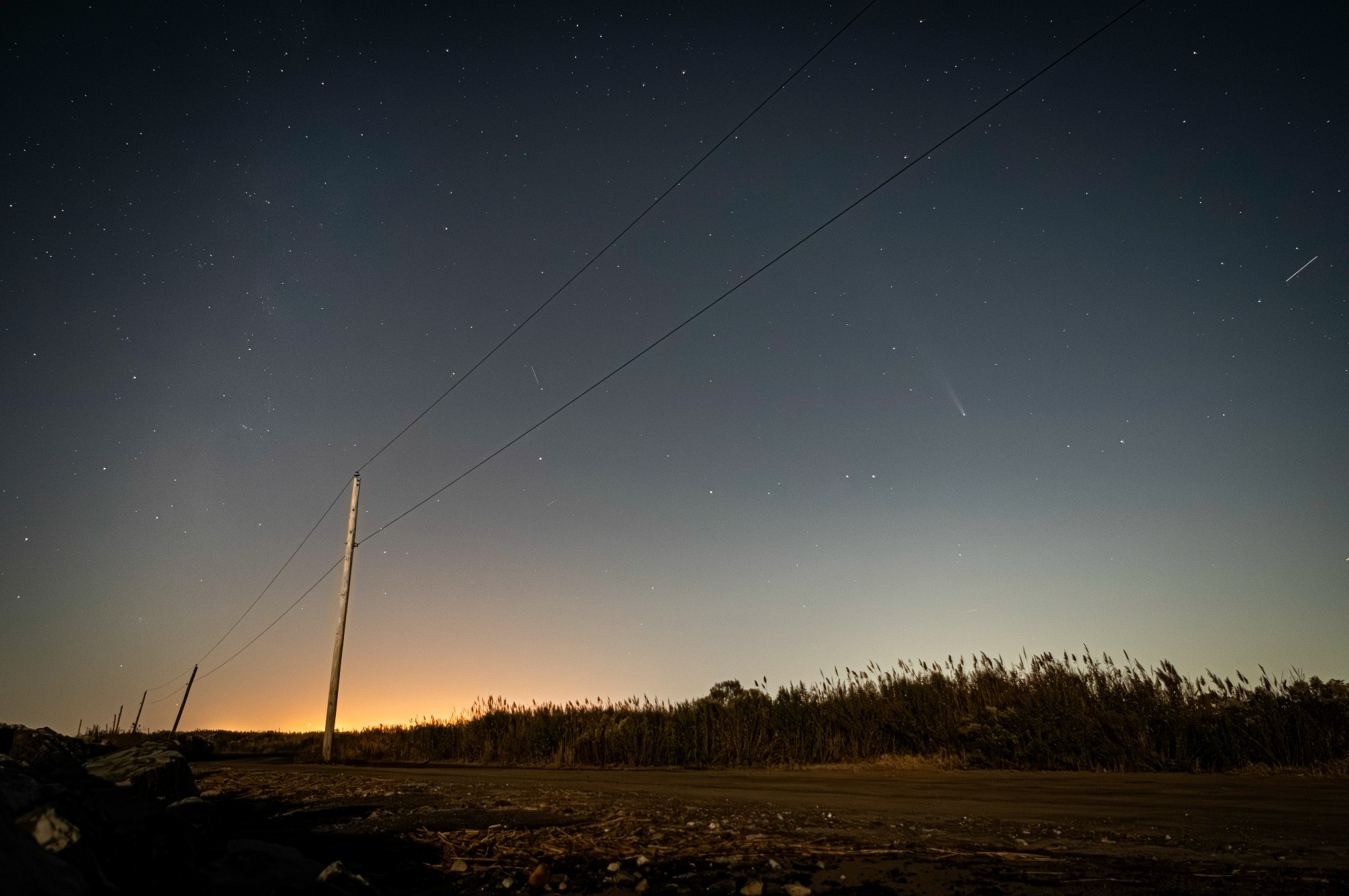 The fading comet is seen at right over cattails and beneath a telephone wire. Stars and the milky way fill the sky. There's a red glow on the horizon of this dirt rocky road.