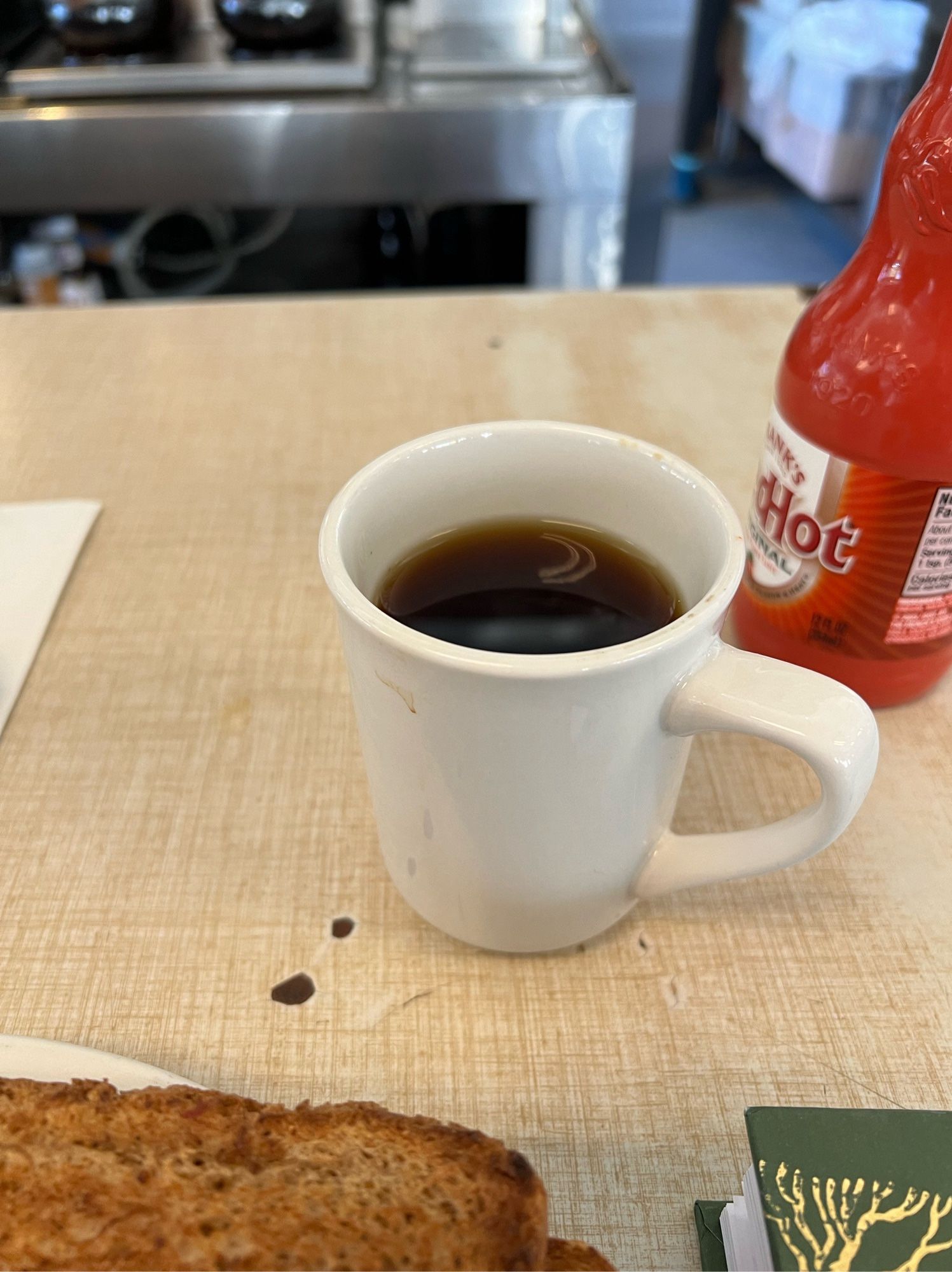 Photo oh a coffee cup reflecting light in a crescent shape with toast, hot sauce and the green cover of the book I’m reading in West Side Diner in Providence RI