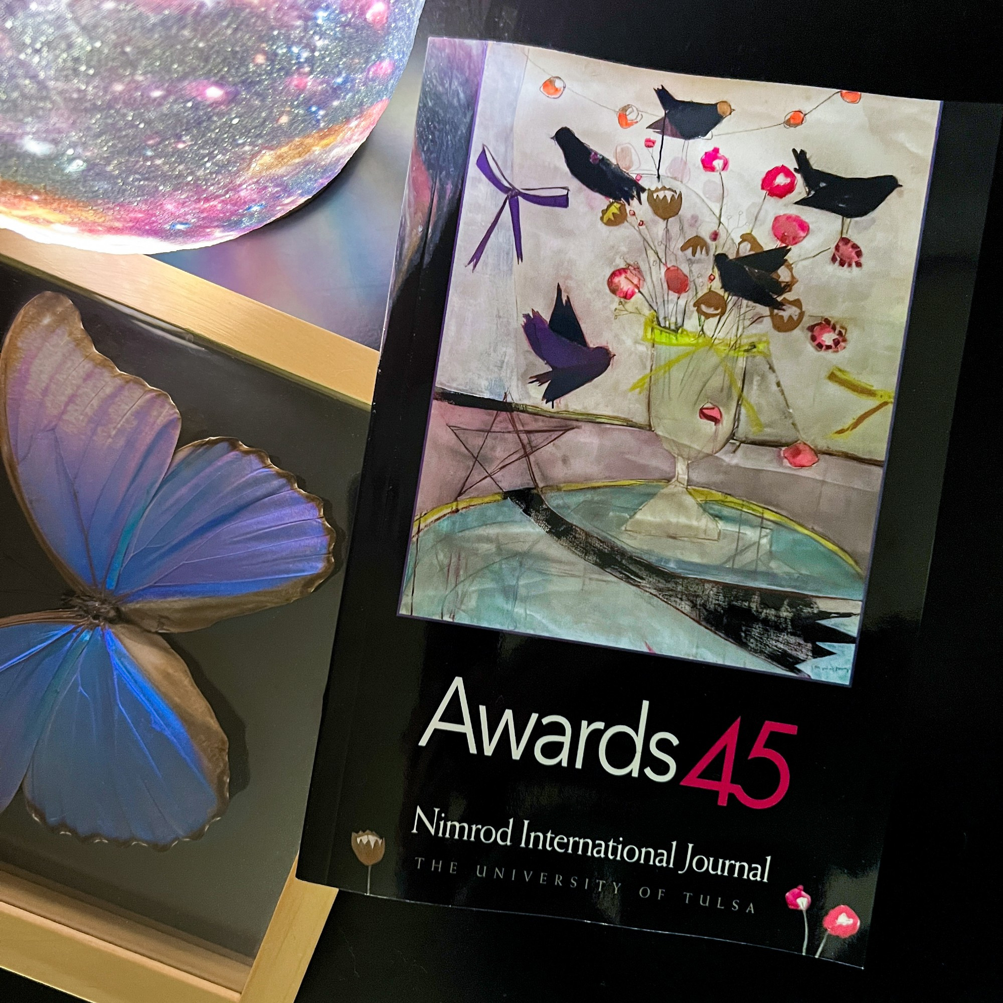 Picture of Nimrod International Journal Awards 45 Issue, with black birds on the cover. Pictured lying on a table with a blue butterfly in a frame and a globe light.