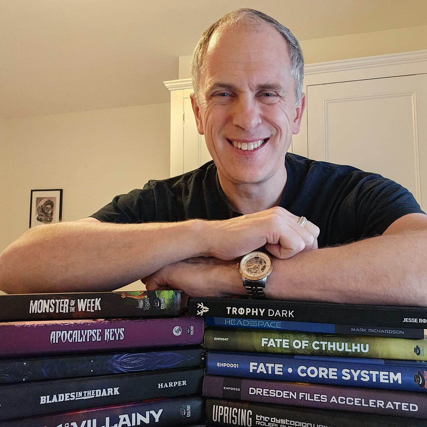 Photo of author with a pile of TTRPGs:
Monster of the Week, Apocalypse Keys, Girls by Moonlight, Blades in the Dark,  Scum & Villainy,  Trophy Dark,  Fate of Cthulhu,  Fate, Dresden Files Accelerated,  Uprising.
In the background: frame with the drawing of a astronaut helmet with a skull in it.