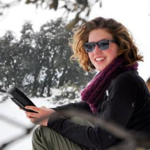 Here, Dr. Mairin Deith (a white woman with short, curly, light brown hair) is sitting in a snowy field with a bulky tablet in her hands. She is smiling at the camera with sunglasses on her face and is wearing a maroon infinity scarf, black jacked and olive green pants. In the background there are dark leafless trees.