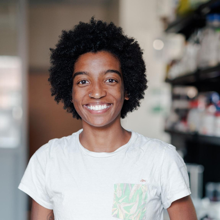 Here, Dr. Peyton Thomas (a black woman, with short curly hair and a silver hoop nose ring on the right side of her nose) is smiling wide at the camera in a white Patagonia t-shirt. She is in focus in the photo and the background is an out of focus lab setting.