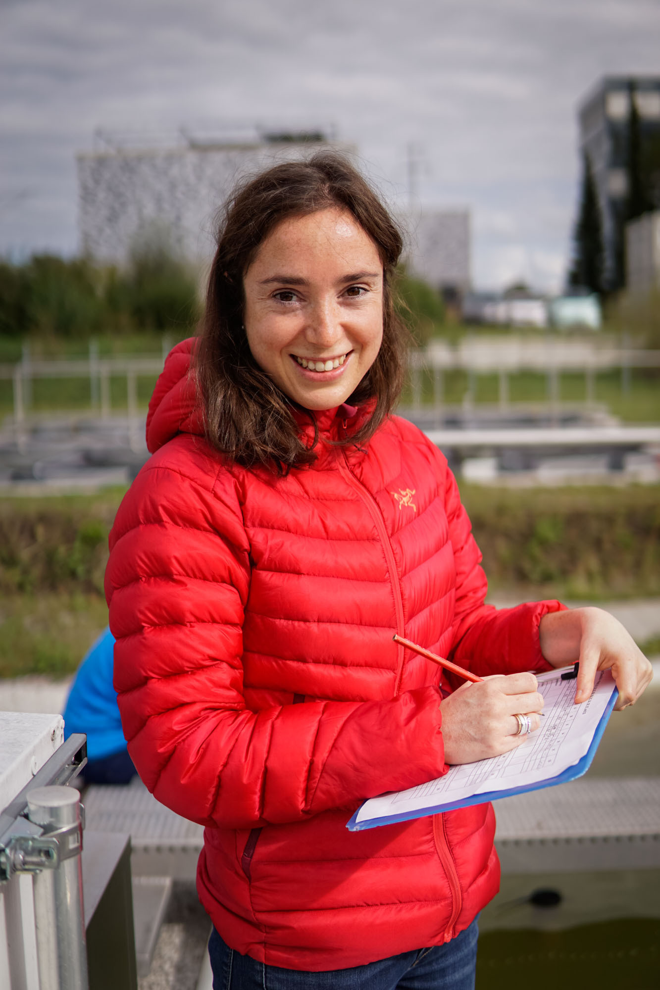 Here, Dr. Joey Bernhardt (a white-passing woman with short brown hair) is standing on a boat (or a metal platform near the water) in a bright red puffy jacket with a clipboard in her hand and is smiling at the camera.