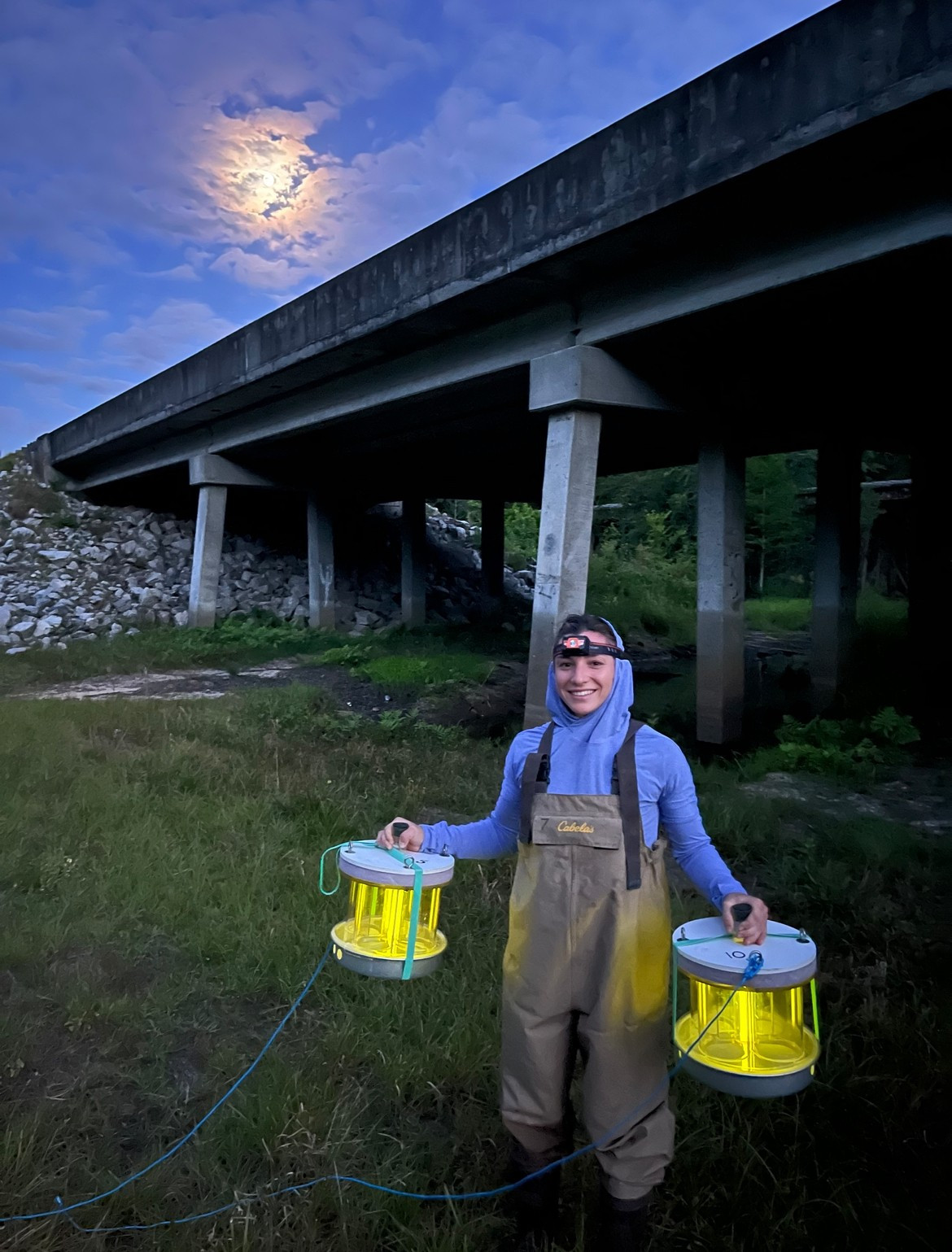 Here Bridgette is pictured in front of a bridge holding lit up light traps  in either hand at twilight. She is wearing a headlamp and Cabelas waders with a periwinkle long sleeve hooded sun shirt.