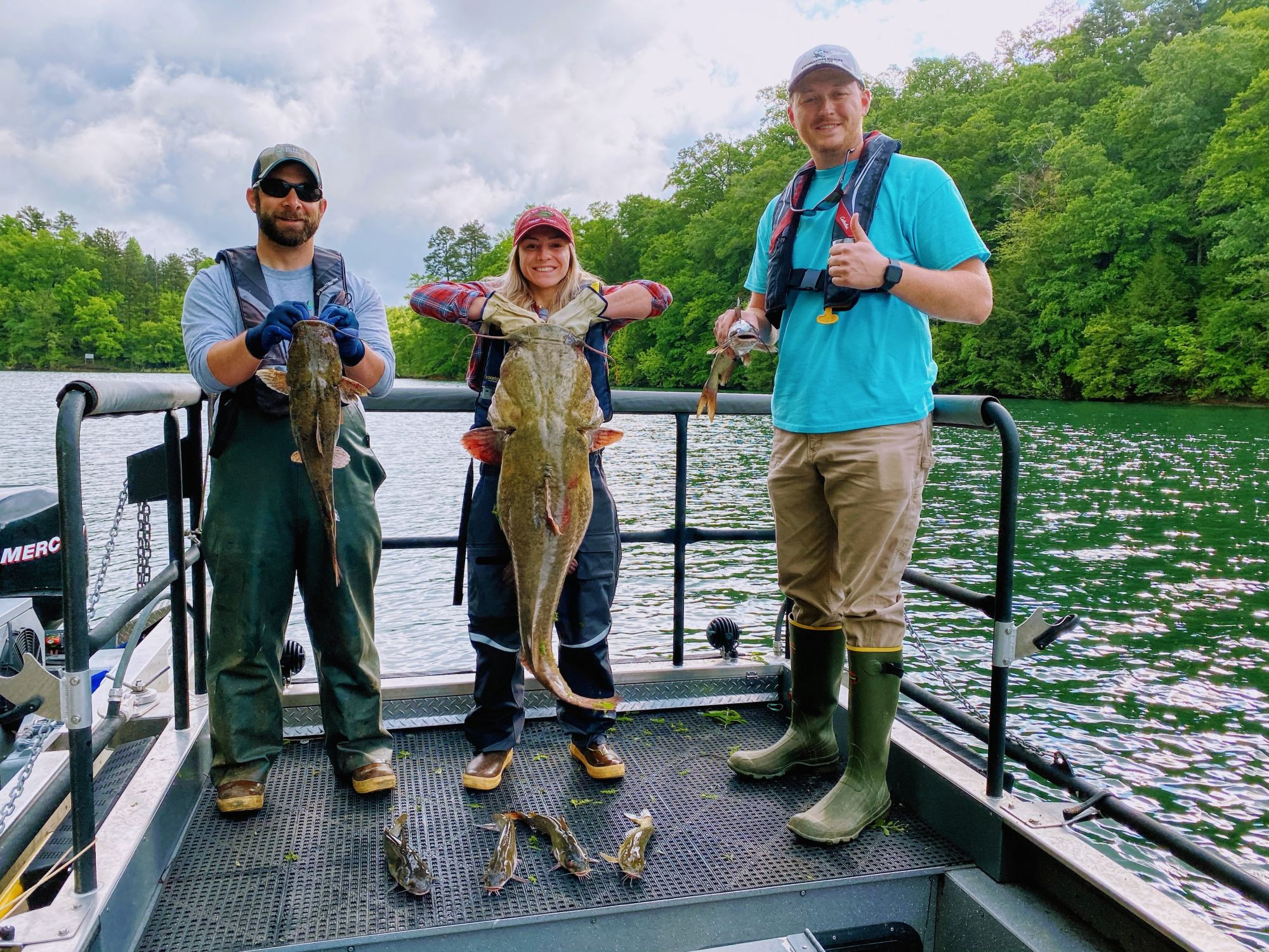 Here Bridgette (an ~5ft tall woman with mid-length blonde hair) is holding a massive catfish that is about 3/4 the size of her in a boat in the river with two men on either side of her (both of which are holding much smaller catfish).