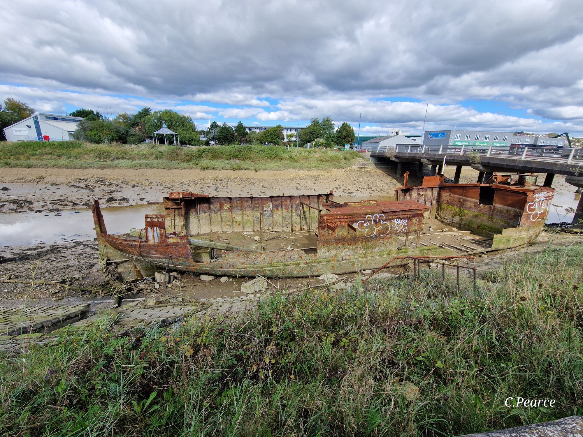 Photo of the boat's port side. It's laying on the river bank at low tide, so it's in the mud. The photo is taken from the main ring road around Newhaven, and the bridge to Denton Island can be seen on the right. What would have been wood is gone; the metal is rusted.