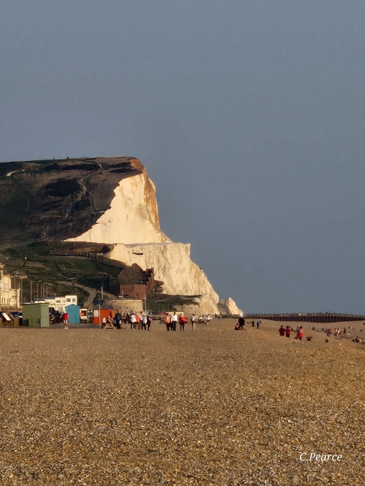 Photo taken towards the east along the shingle beach, showing Seaford Head, the round Martello Tower at the base, and white and green beach huts along the left side. There are people in front of the huts, as well as down towards the water. The sky is blue and hazy.