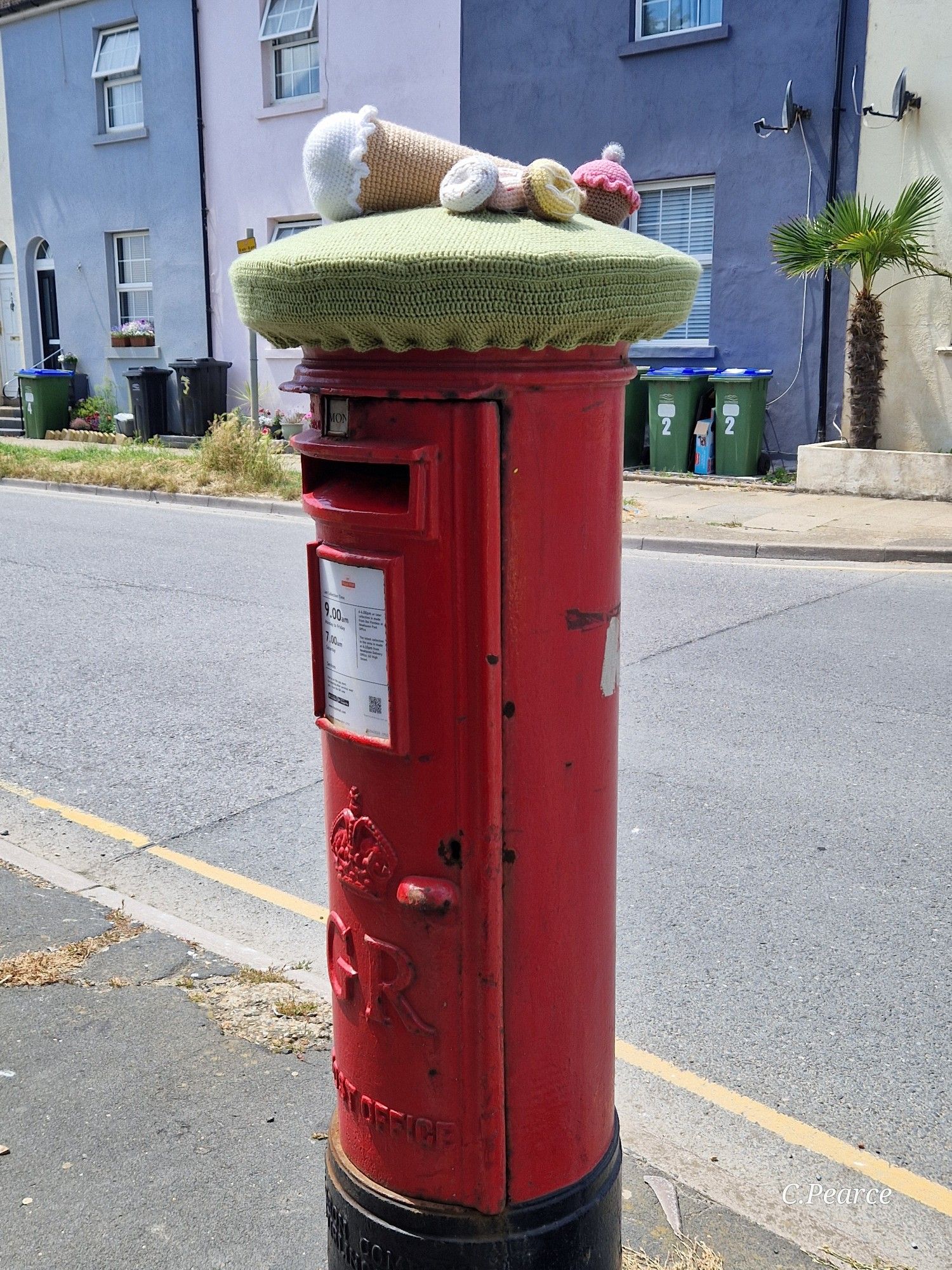 Photo showing a red cylindrical post box on a street with a colourful painted row of houses in the background. The post box has a green knitted hat, a knitted vanilla ice cream cone, two frosted donuts, one with white icing and the other yellow icing, and a pink frosted cupcake.