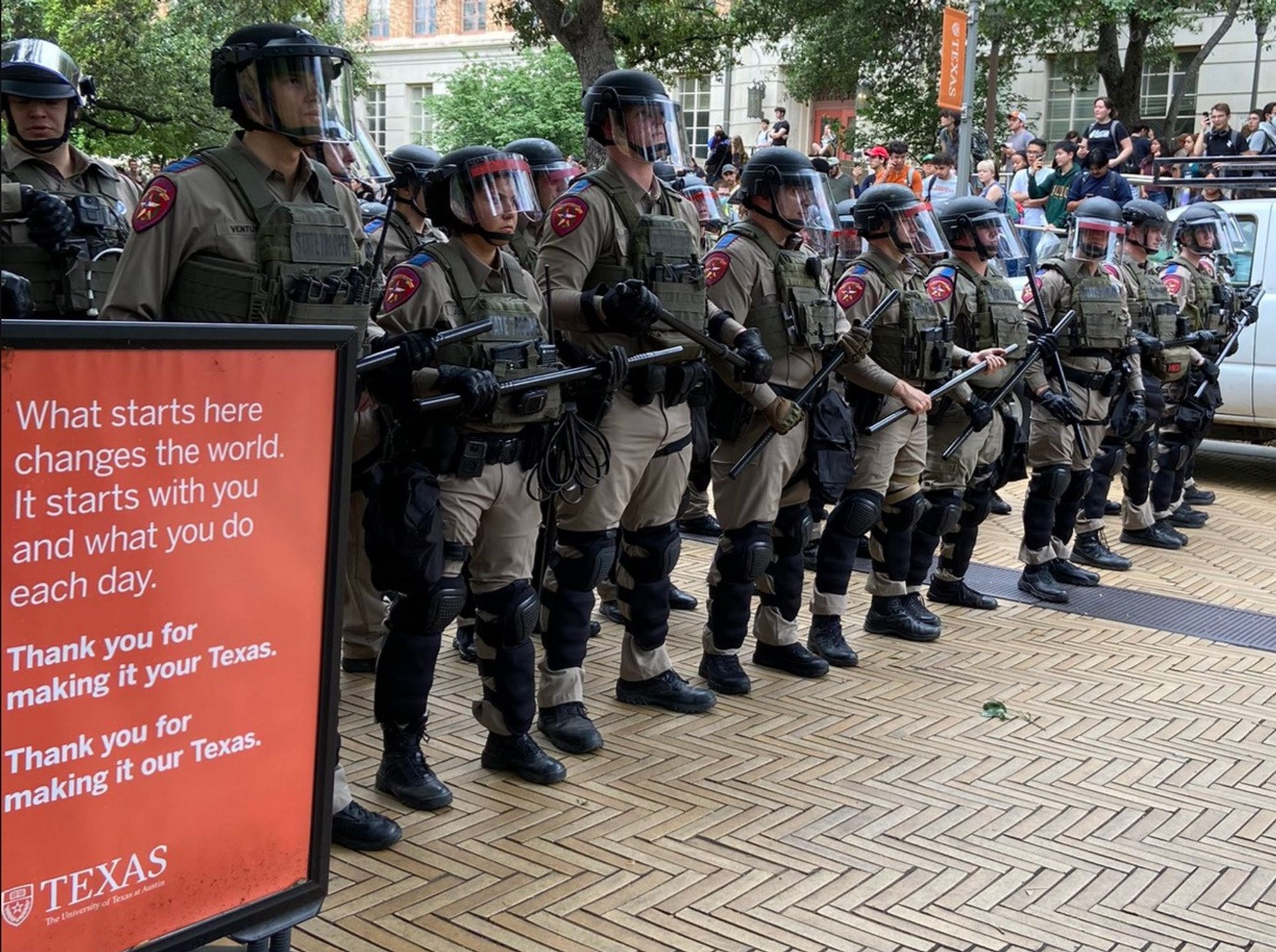 Riot police stand next to university sign that reads, "What starts here changes the world. It starts with you and what you do each day." From Austin UT.