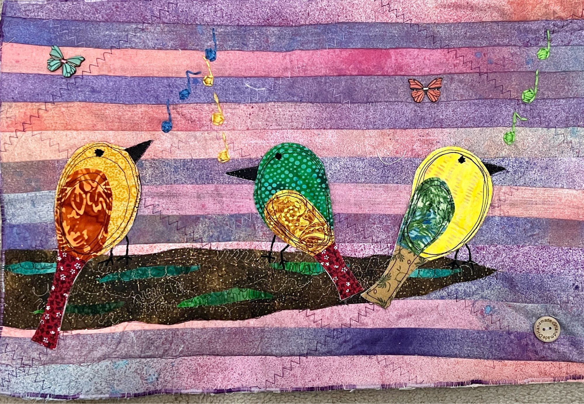 3 birds created with fabric resting on a large brown limb. Their feet are hand stitched with music notes drifting above them. Two small butterfly buttons are attached in the sky of alternating strips of hand dyed purples and pinks