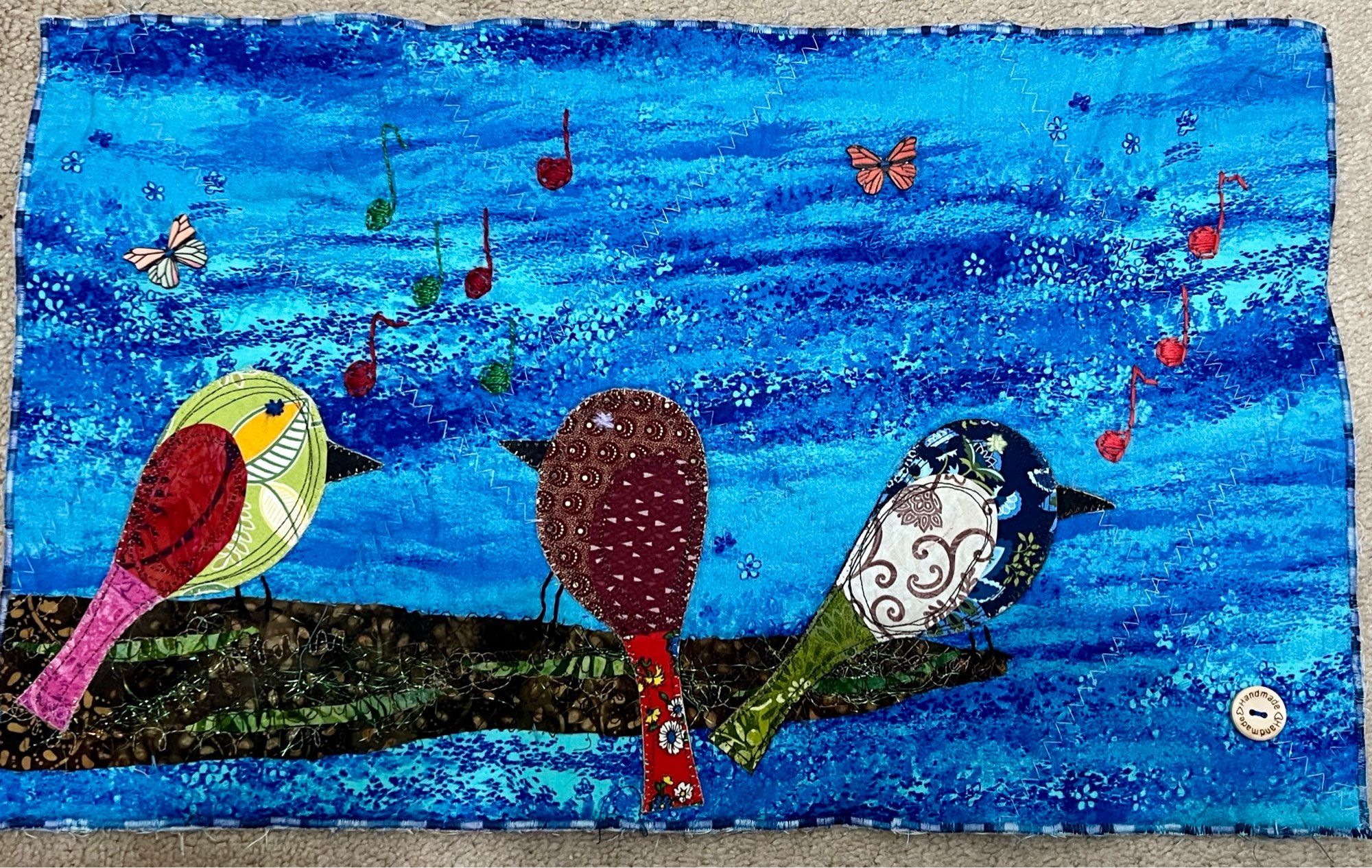 3 birds created with fabric resting on a large brown limb. Their feet are hand stitched with music notes drifting above them. Two small butterfly buttons are attached in the sky of wispy blues scattered with tiny flowers.