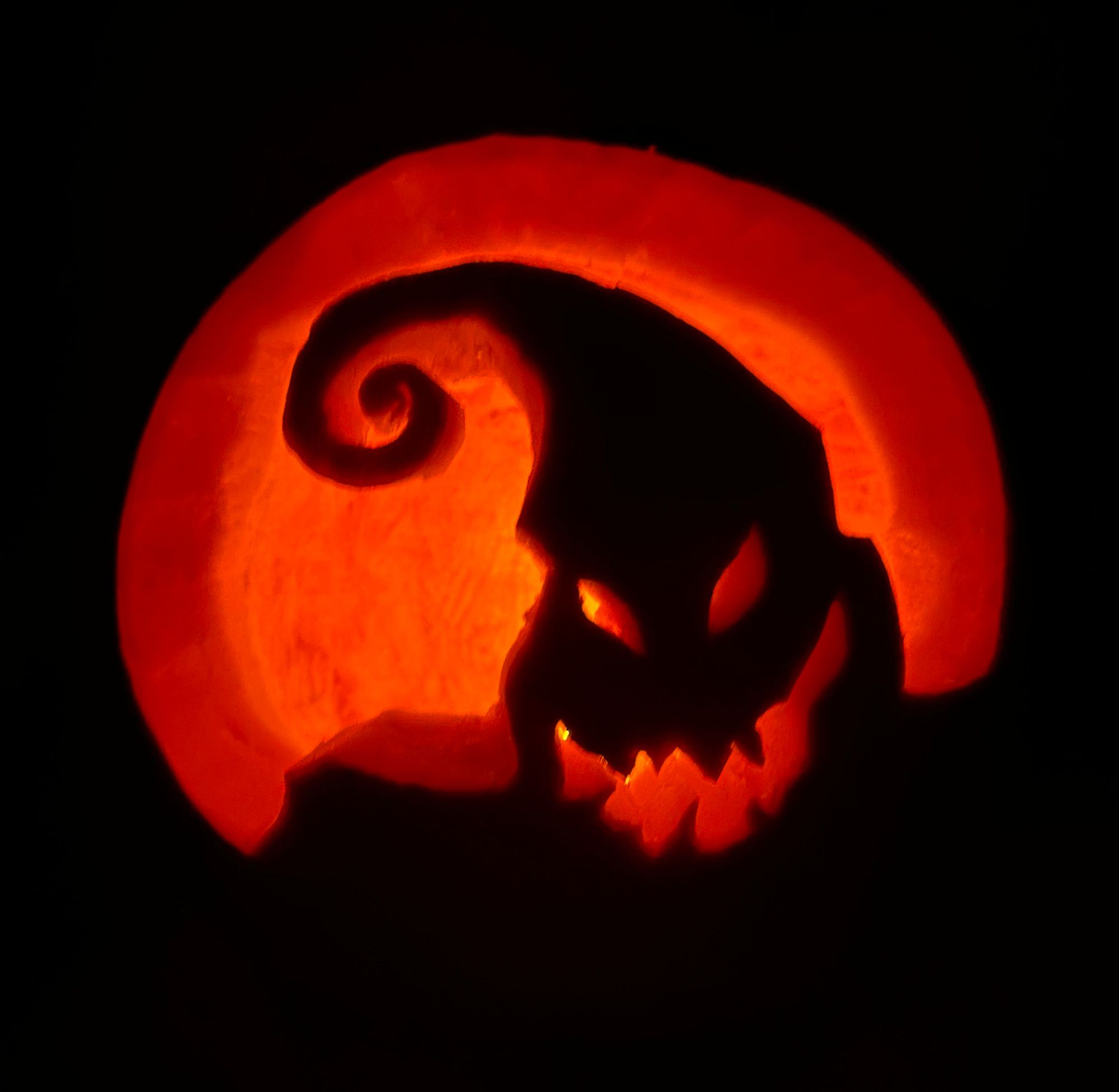 The silhouette of Mr. Oogie Boogie (from The Nightmare Before Christmas) against the moon - carved into a pumpkin, and lit from behind. The moon, his eyes, and his mouth are glowing various shades of orange and yellow.