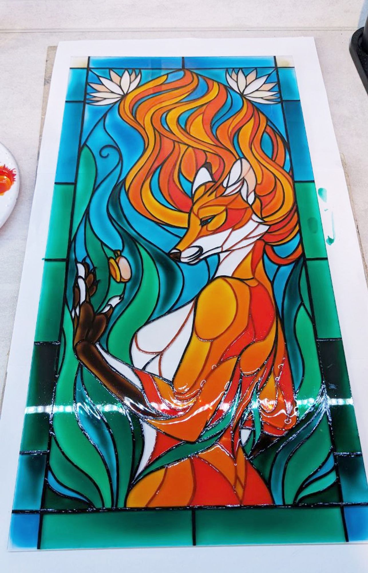 Photo of an anthropomorphic vixen painted on glass, fashioned like a stained glass window. The vixen is holding an amulet and appears to be underwater, judging from all the blue and green flowy segments of the piece.