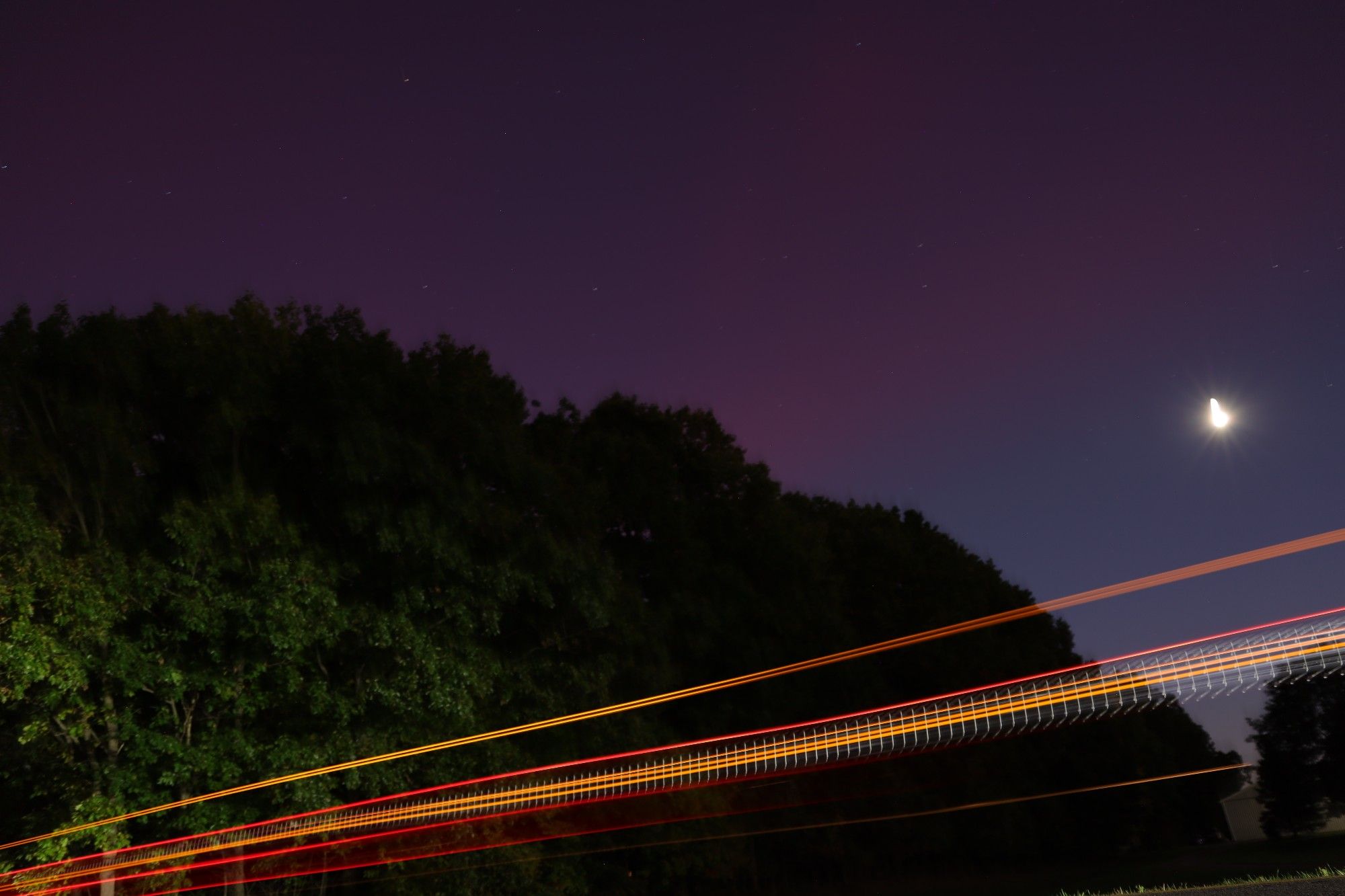 dark treeline with the moon to the right. A pink arc extends from the top left, forming a line near the moon. In the foreground, the red brakelights of a truck extend in a dazzling streak across the entire photo