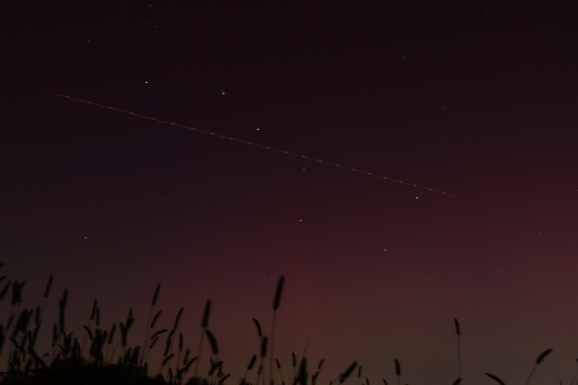 plane streak the big dipper in a pink-lit sky; the line is dashed red and white as the plane's lights blink