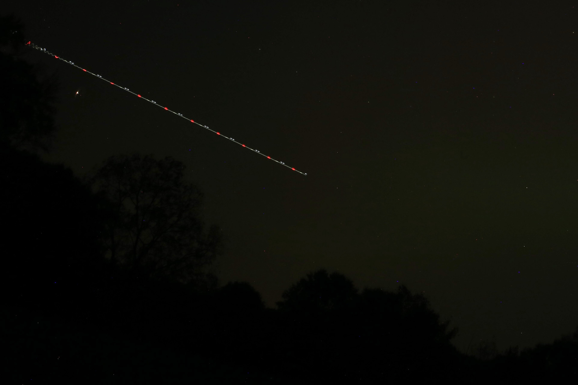 plane streak over a murky dark green sky; the line is dashed red and white as the plane's lights blink