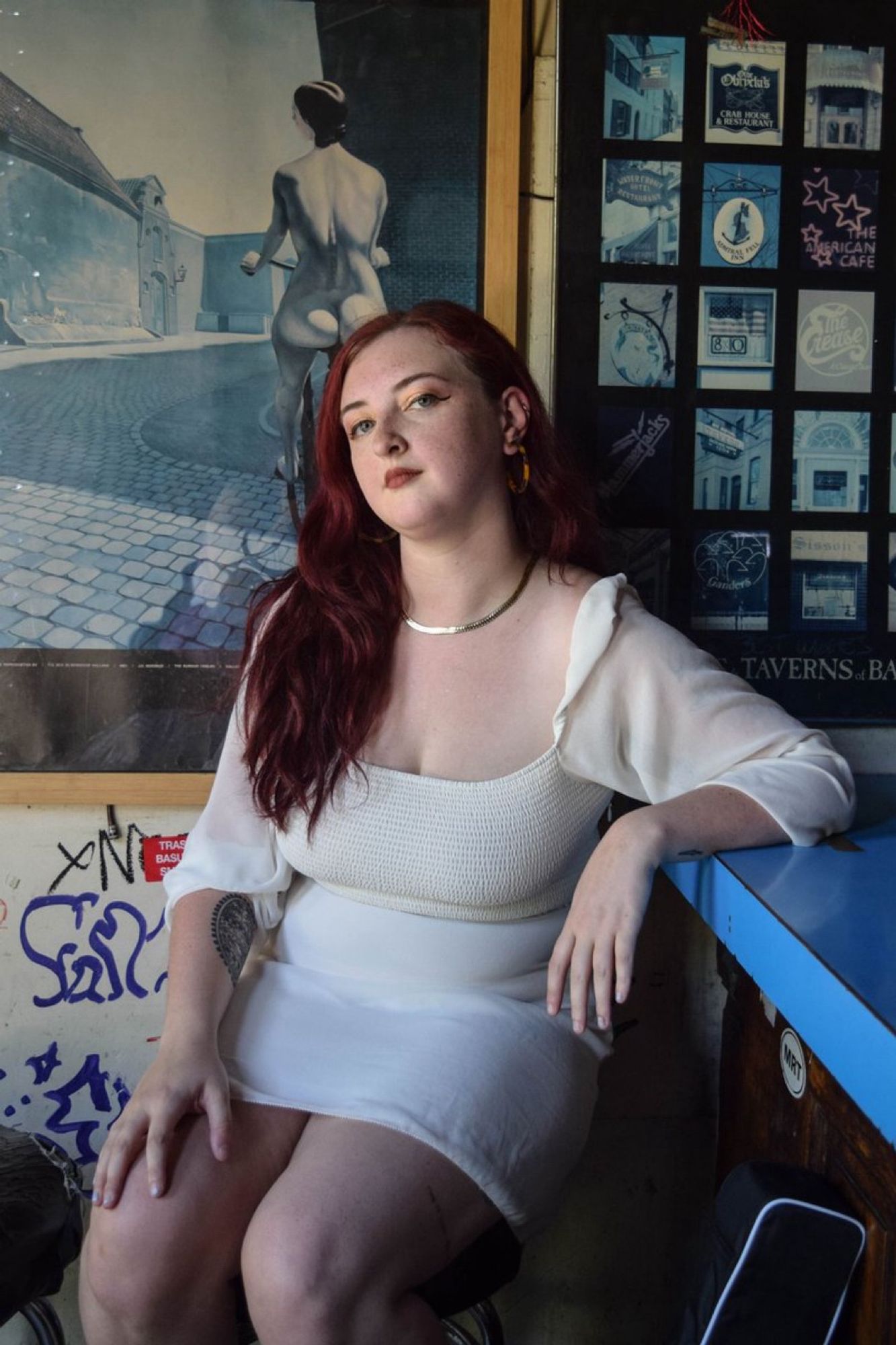 Alt text: nat raum, a white nonbinary person with red hair, sits on a barstool at the Mt. Royal Tavern wearing a white dress and their grandmother's gold chain.