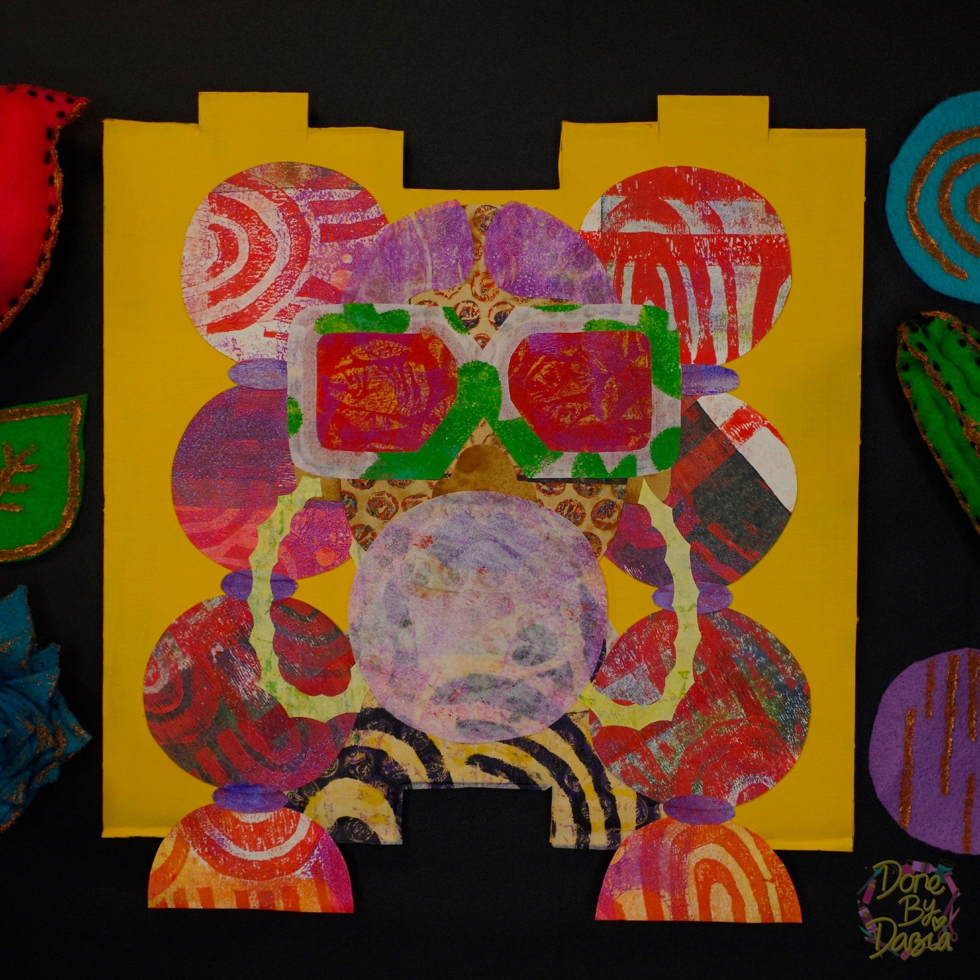 A portrait made with boldly colored and patterned paper, framed by felt plants. A girl wearing large, rose-colored glasses blows a big bubble of gum. Her hair is up in high pigtails, with ties down the length sectioning off large bubbles of hair.