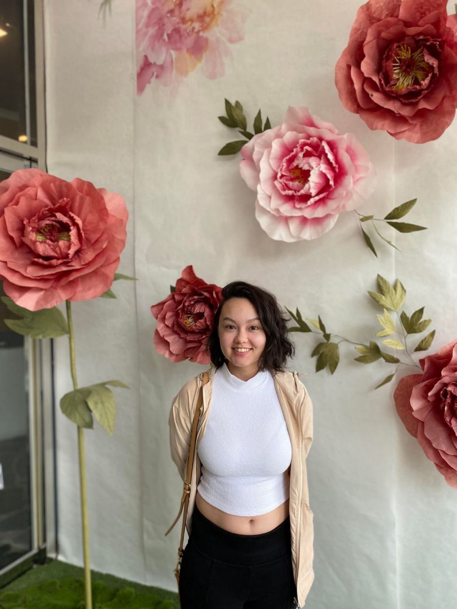 An Asian woman with short brown hair stands in front of a white background with large pink paper flowers in a white shirt and smiles.