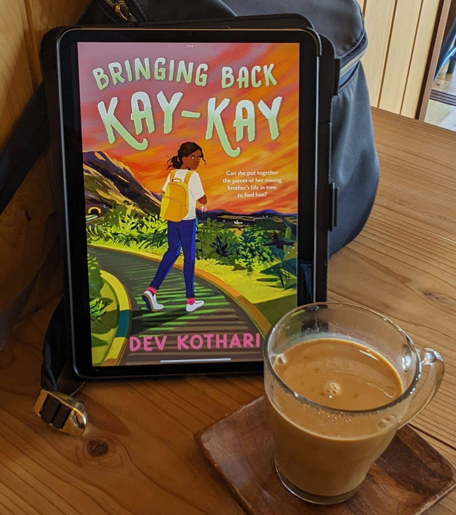 Book cover of Bringing Back Kay-Kay shown on iPad, propped on a blue backpack. A cup of chai in the foreground (slightly to the right). It's served in a clear glass cup. Everything is placed on a light brown wooden table. 1:1 image