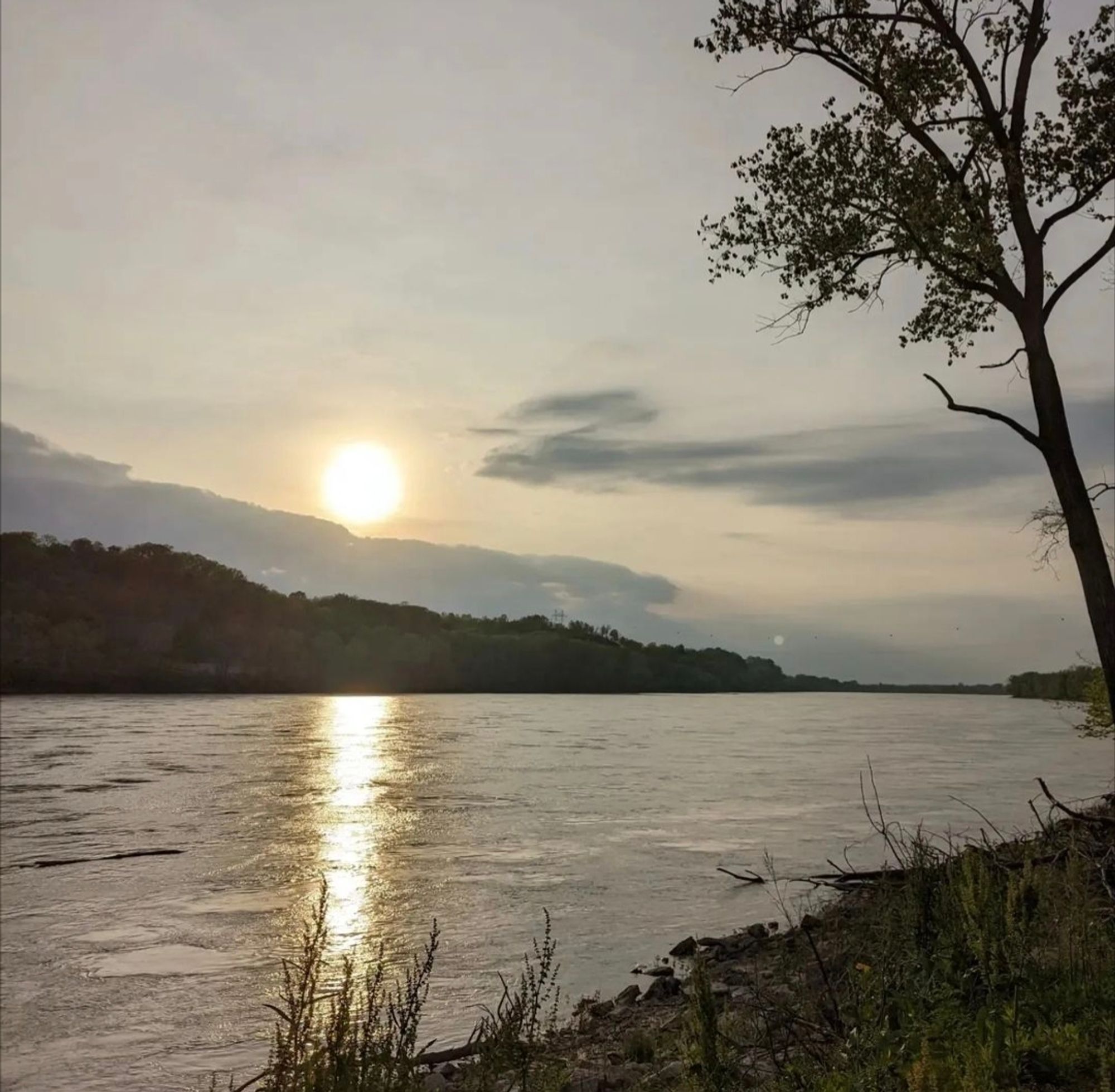 A wide, slow moving river near sunset. The river is hemmed by forested hills.