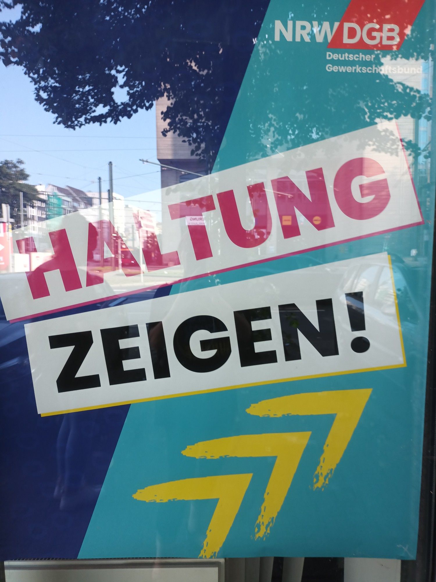 Plakat vom DGB NRW. Blau und türkis mit gelben Pfeilen und der Aufschrift in schwarz und pink Haltung zeigen.