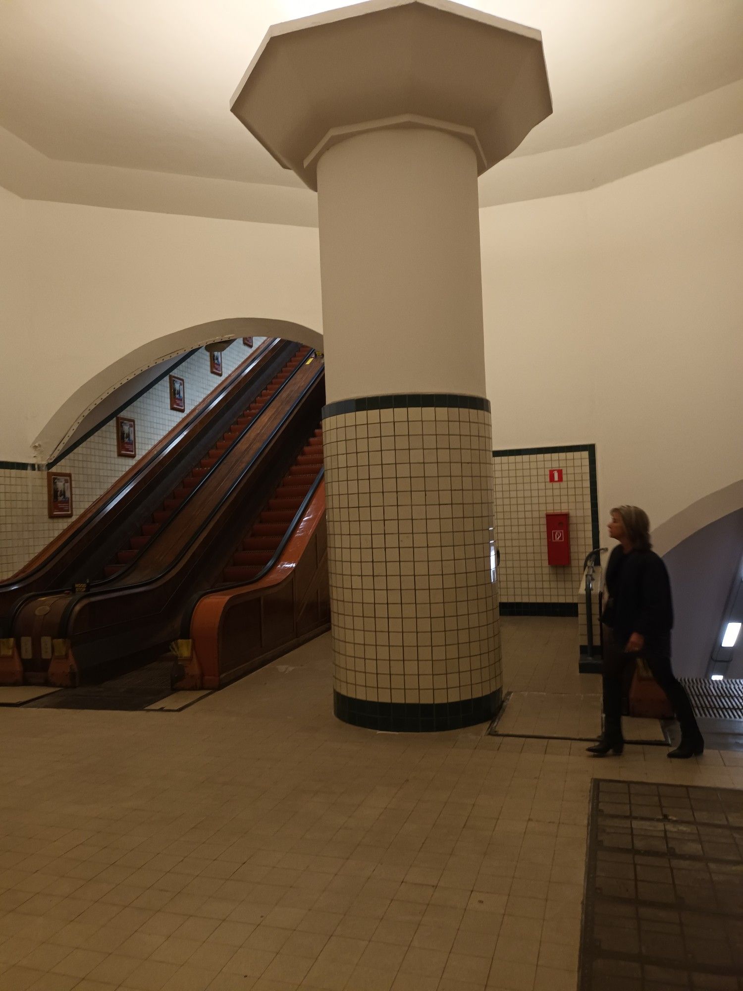 Man sieht eine Rolltreppe, eine Säule mit weissen Fliesen, von rechts läuft eine Person durch das Bild.Die Halle ist Zwischenebene zur nächsten Rolltreppe, die noch eine Ebene tiefer in den Tunnel führt. Man befindet sich in ca 80 m Tiefe unter der Schelde. Antwerpen.