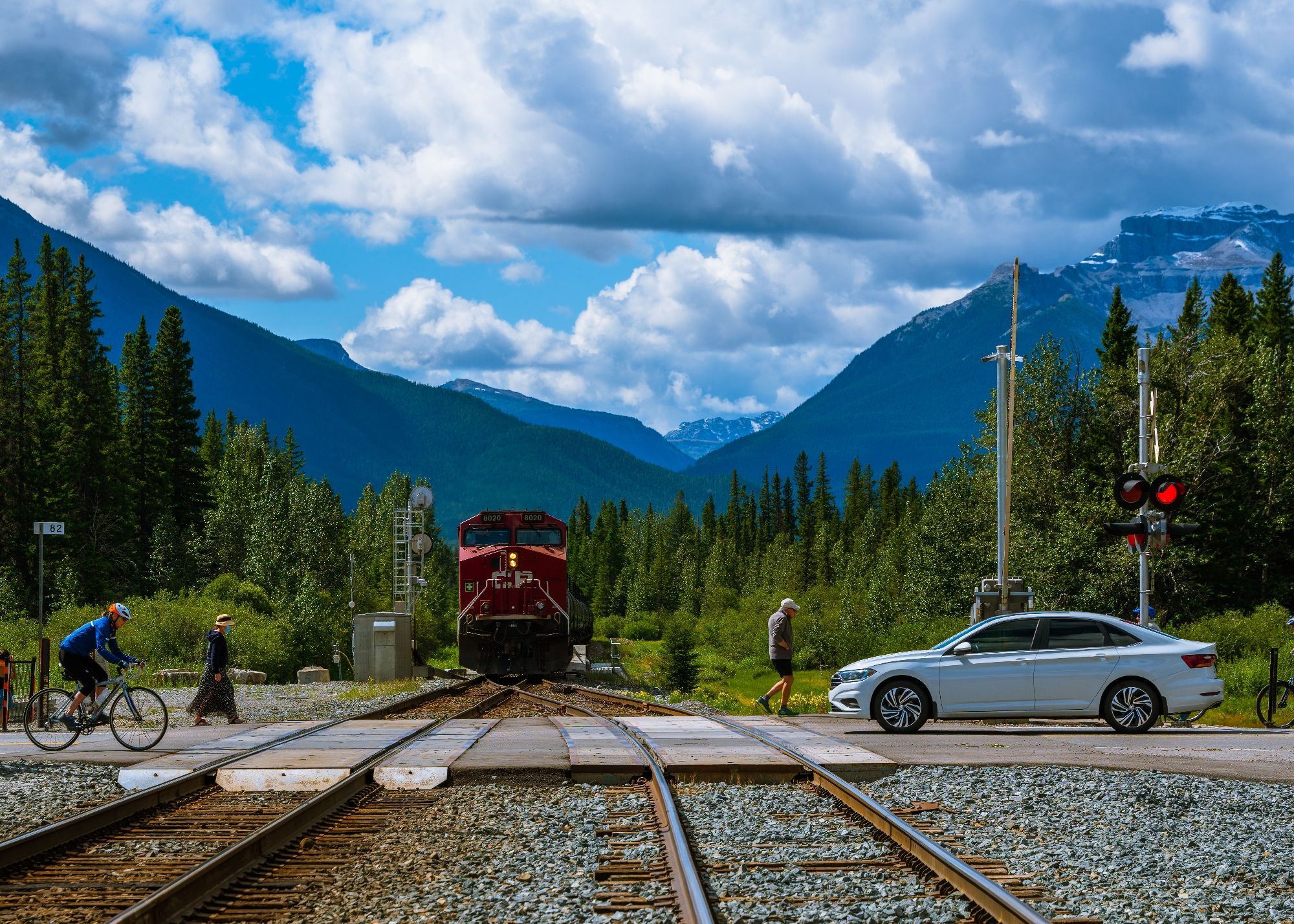 Car, cyclist, and pedestrians crossing rail tracks as the Canadian Pacific iconic red locomotive pulls out of Banff Train Station under partly cloudy skies with Banff National Park pine woods and Sundance Canyon in the backdrop
Captured by Komeil Karimi