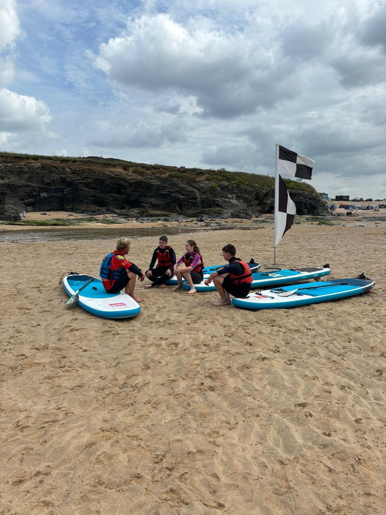 Photo of Emily and two teenagers sitting on paddleboards on the beach listening to a briefing from an instructor.