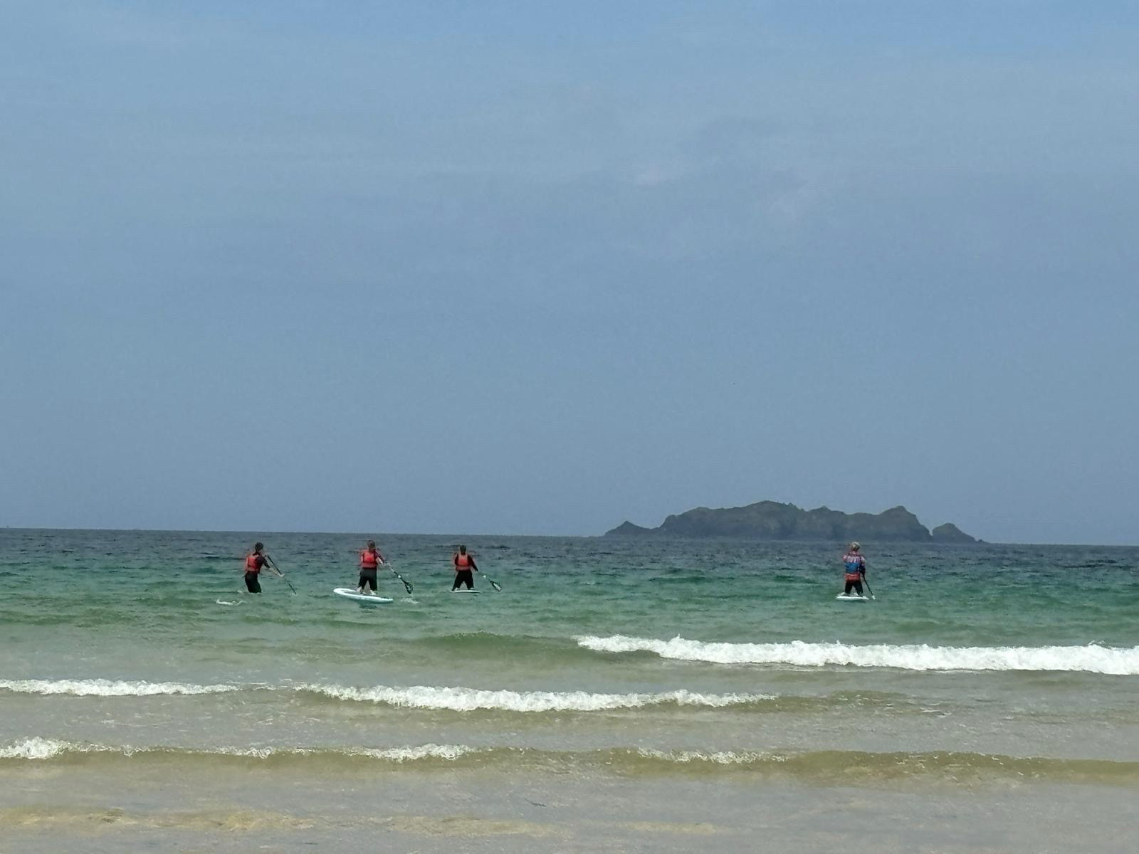 Photo of Emily and two teenagers kneeling on paddleboards out at sea with an instructor. There is an island in the background.