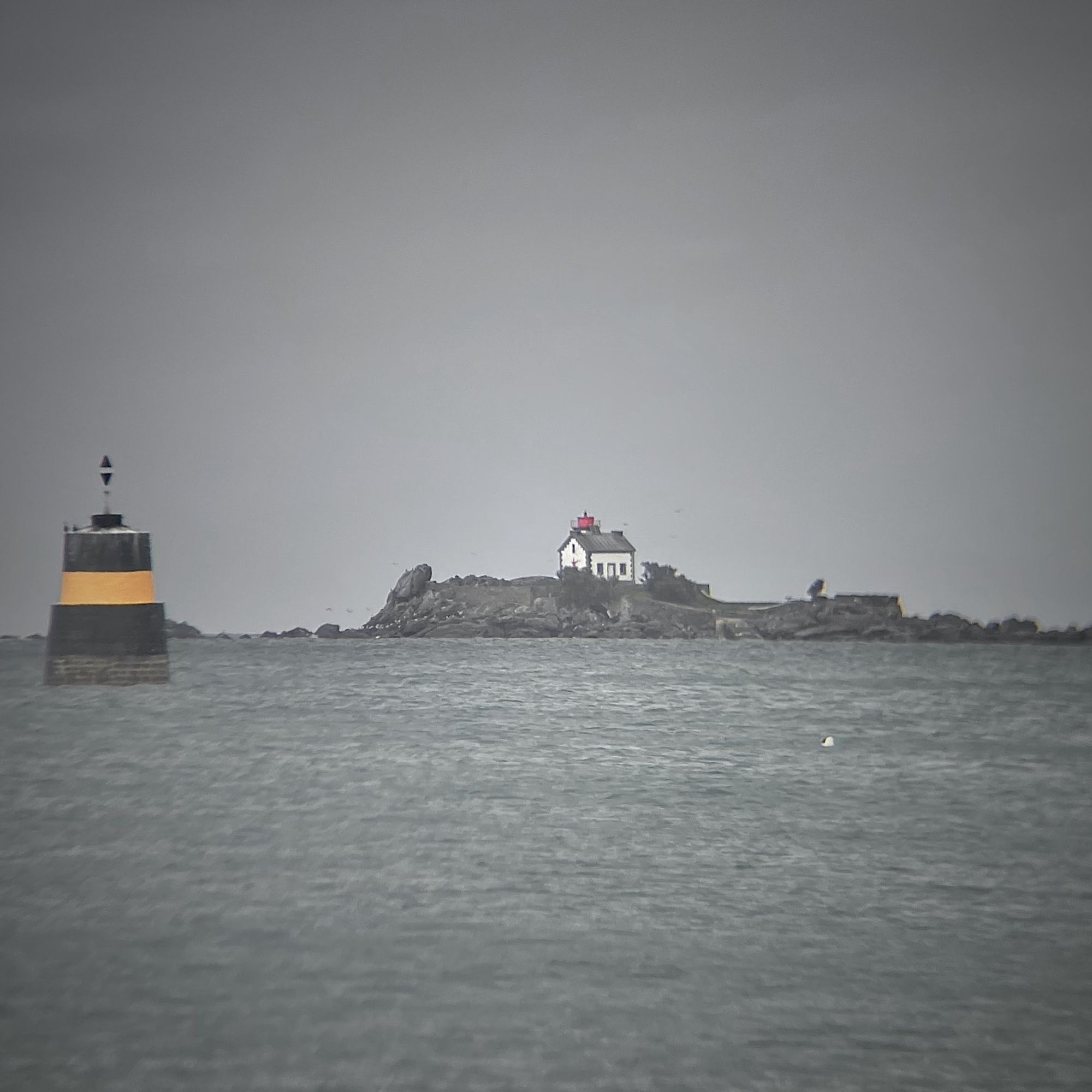 Maison-phare sur un rocher en pleine mer près d'une tourelle noire et jaune avec un ciel gris et une mer grise