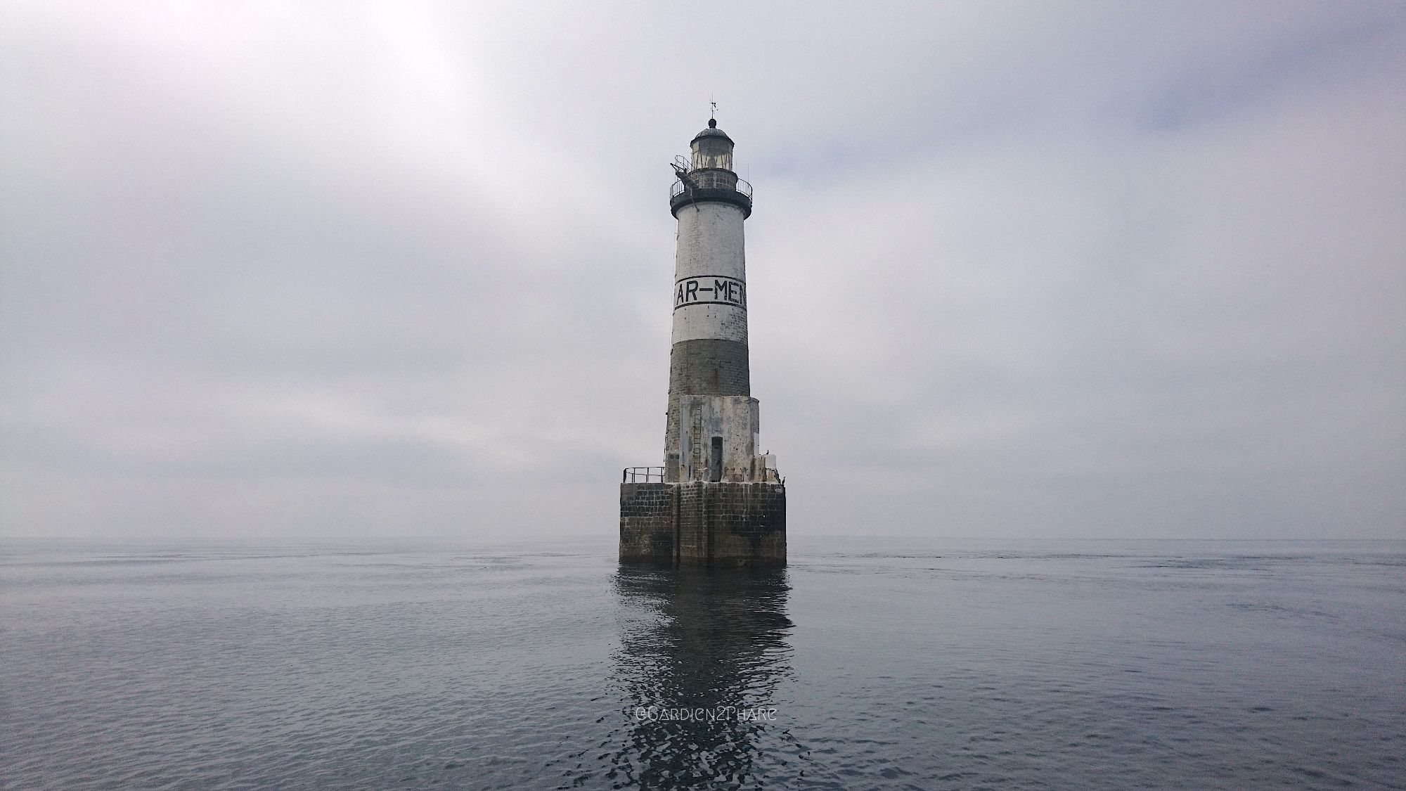 Phare d’Ar-Men, phare blanc et gris construit sur une riche en pleine mer à l’extrémité de la chaussée de Sein photographié ici dans une mer très calme sous un ciel gris