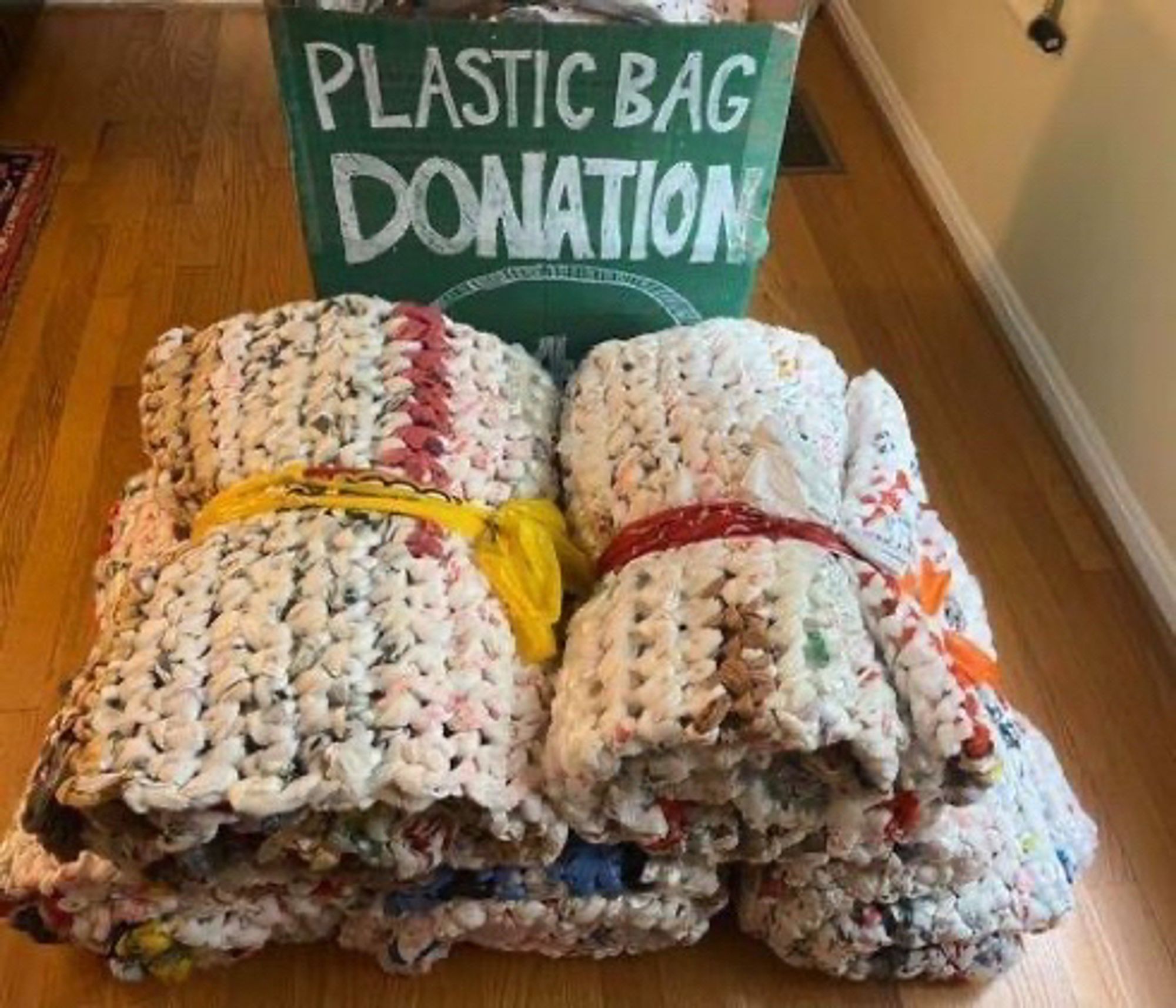 A pile of rolled up sleeping mats made of woven plastic bags sitting in front of a cardboard box with “plastic bag donation” written on it in white