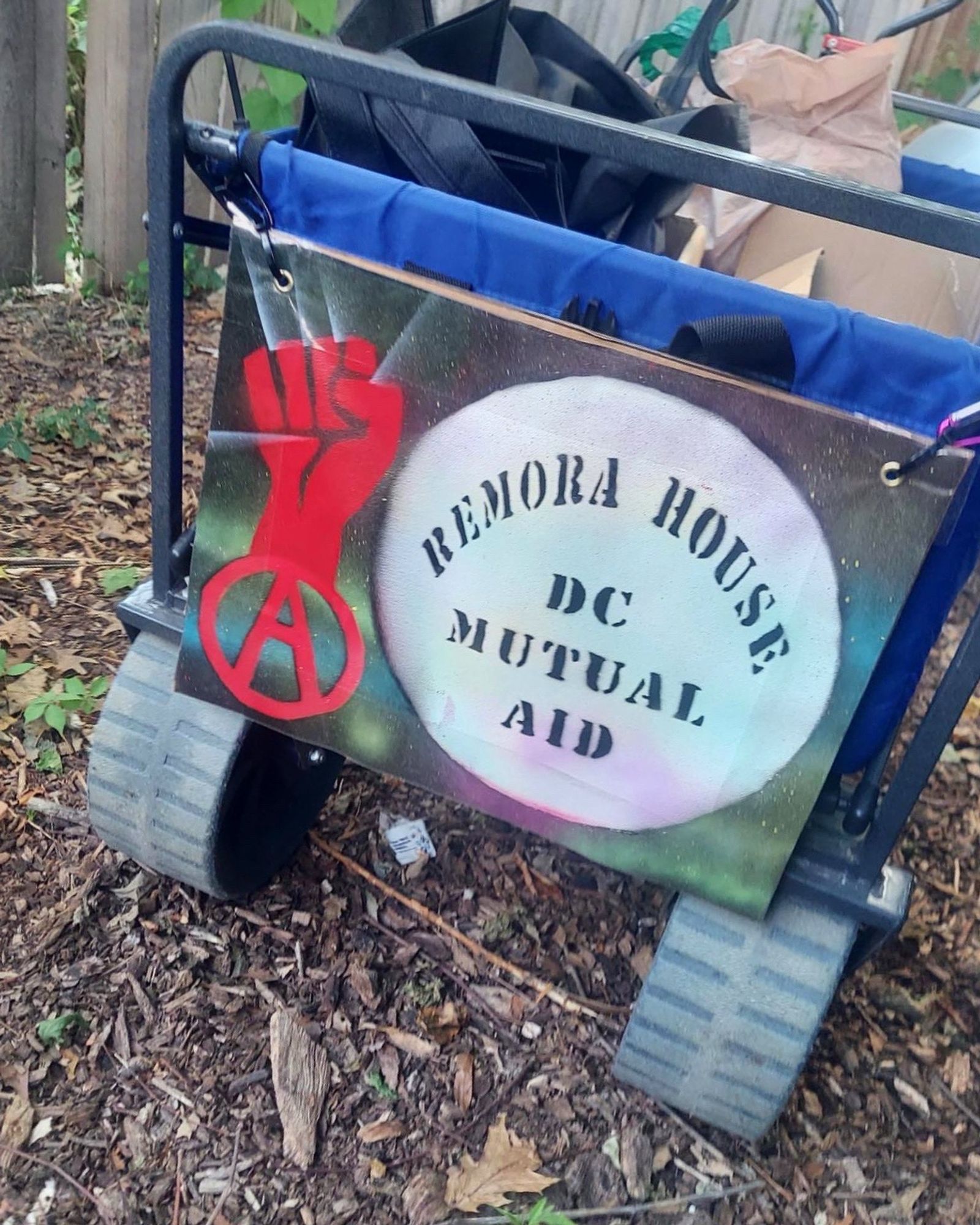 A blue rolling cart on a mulchy ground with a colorful painted sign attached to the front with “remora house dc mutual aid” in a white circle with a red fist above a red anarchy a to the left of it