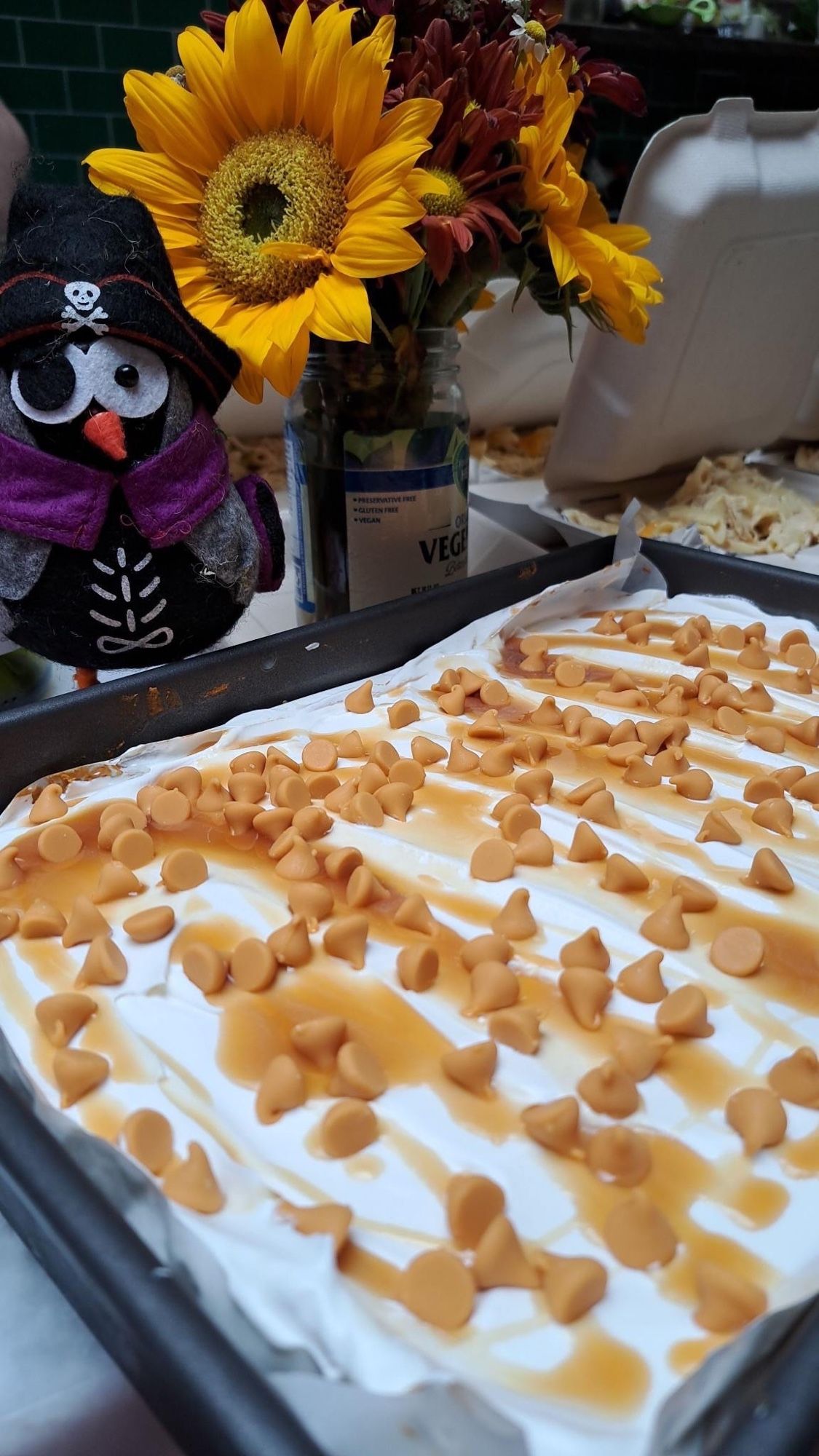 A pan of pumpkin cake topped with whipped cream, drizzles of caramel, and butterscotch chips. Behind the pan is a little felt owl that is dressed up like a skeleton pirate and a cup or orange and yellow flowers.