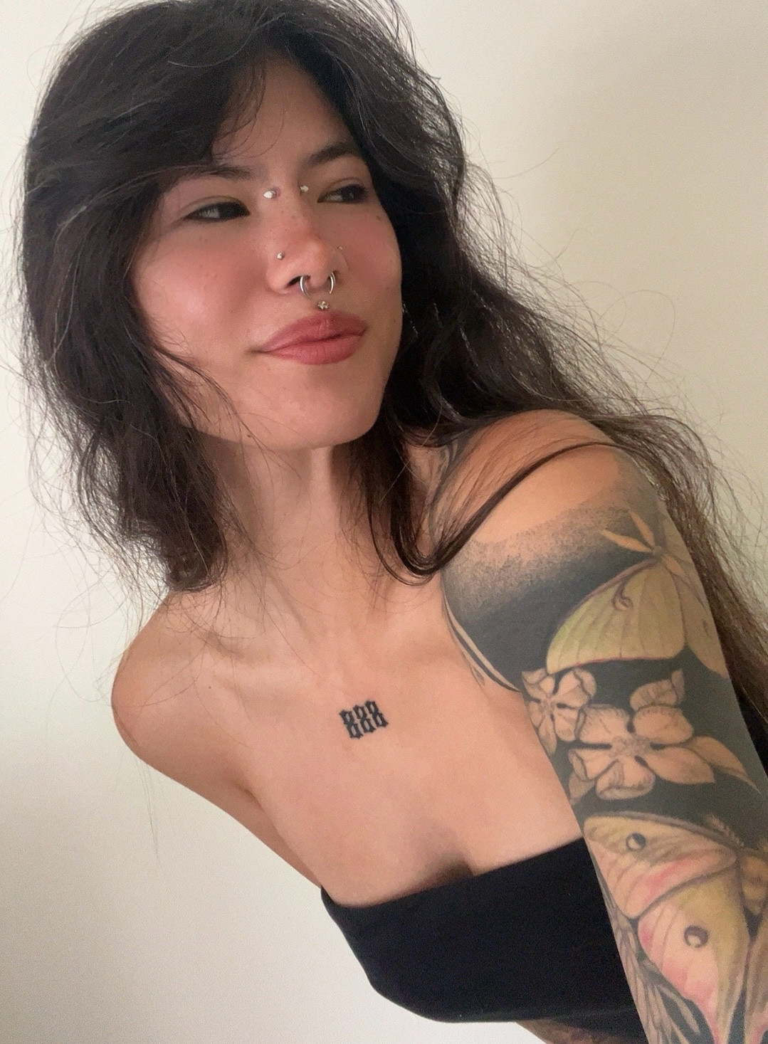 Selfie of HamonCutta looking off to the viewer’s right! She’s wearing a black tube top and has a full blackwork sleeve featuring cherry blossoms and luna months and 888 on her chest. She also has many visible facial piercings. 