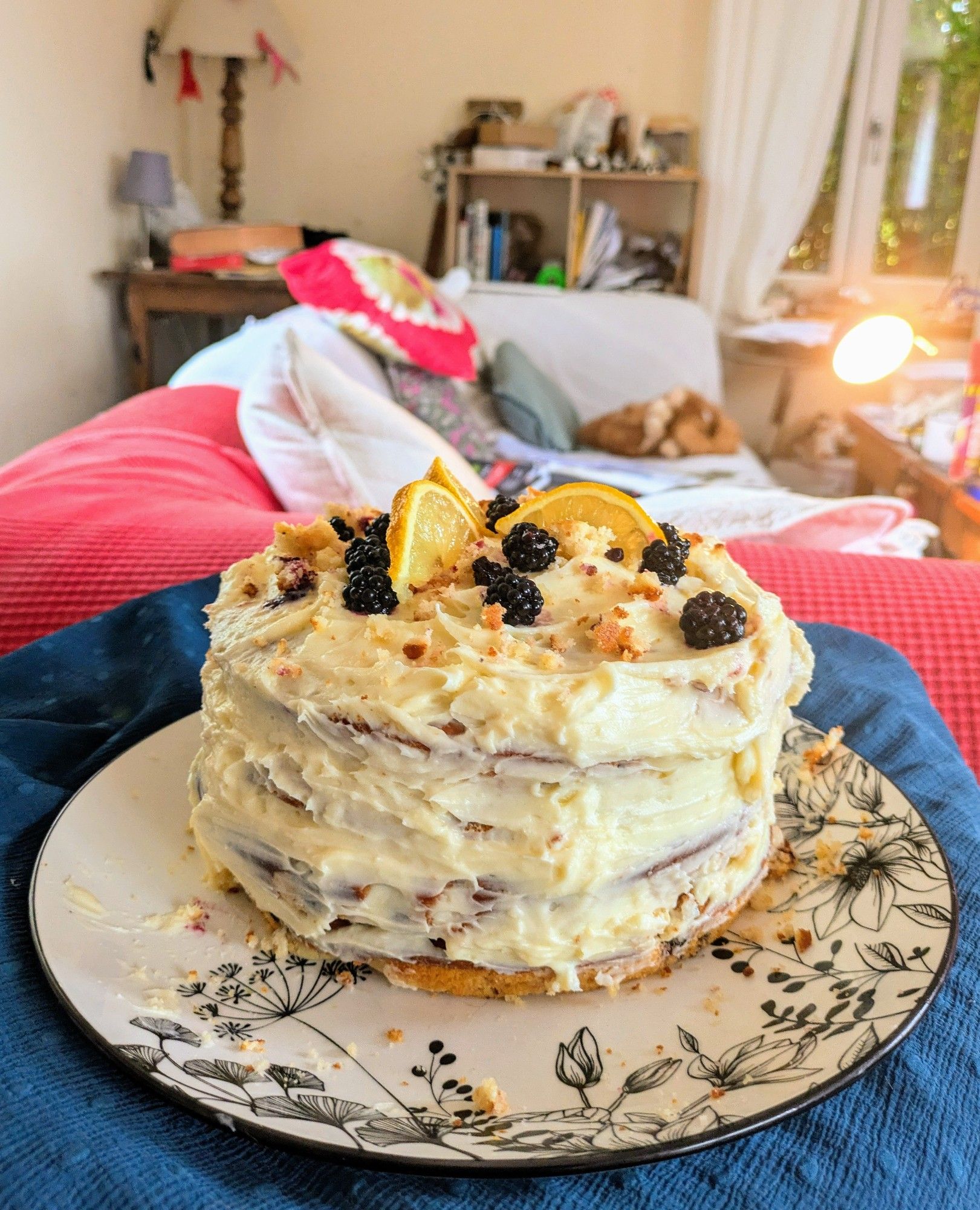 A lemon and blackberry cake with cream cheese frosting topped with blackberries, lemon segments and crumb, on a table