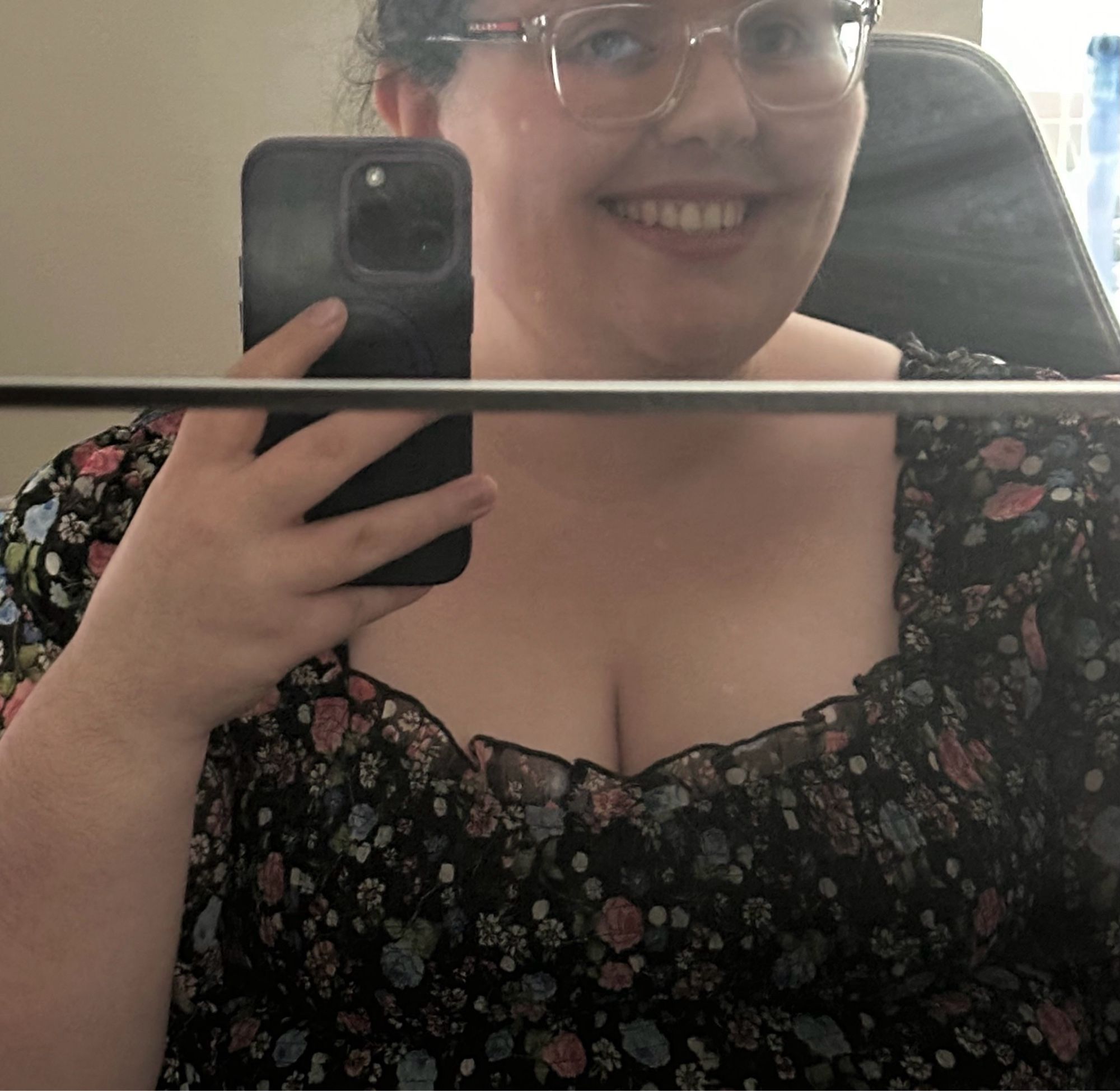 A white woman smiling in a mirror selfie, wearing a low cut floral blouse with prominent cleavage. The work was good, but the view during my presentation was better 😇