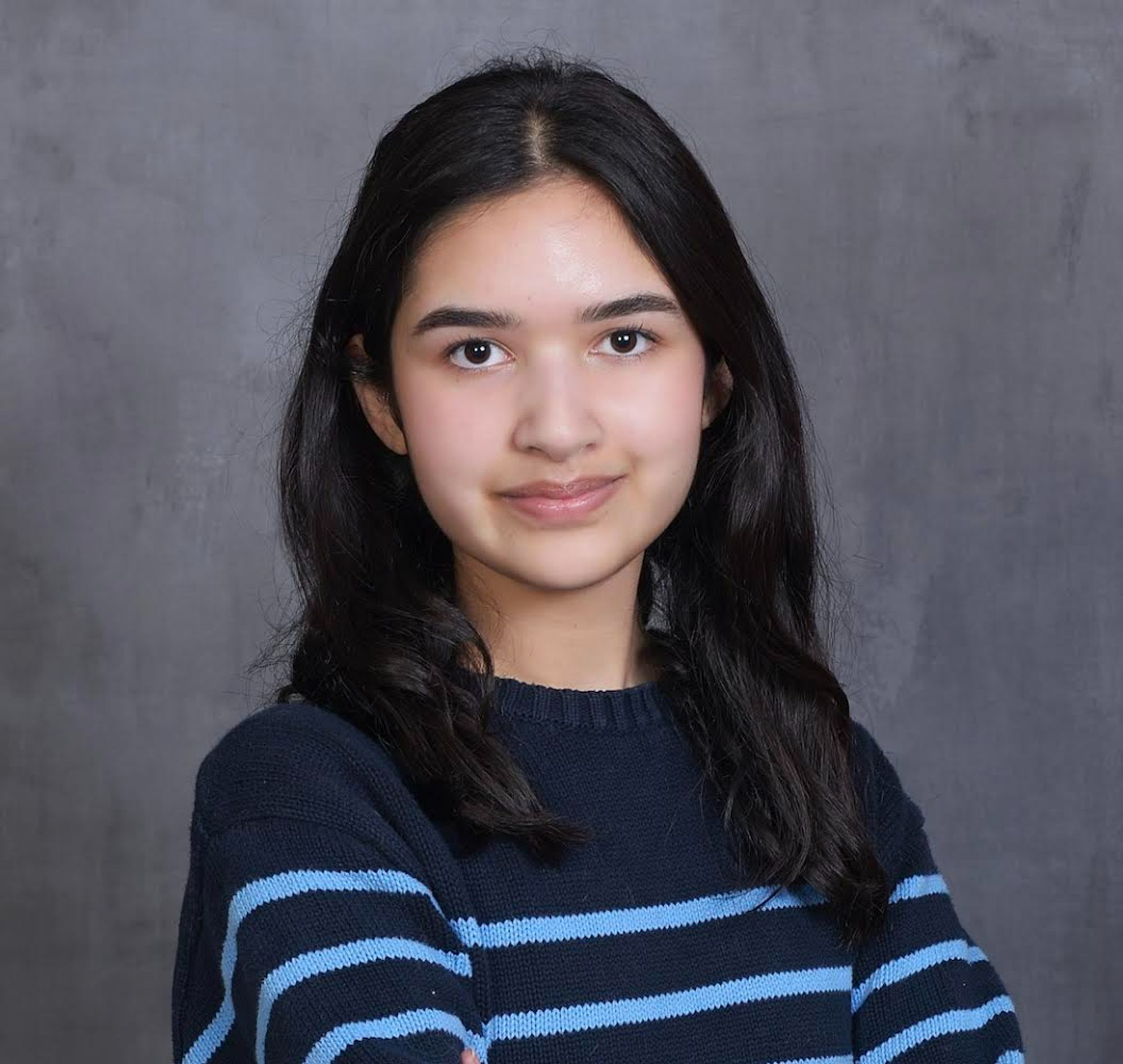 A young lady with dark hair and dark eyes wears a blue striped sweater on a gray background.