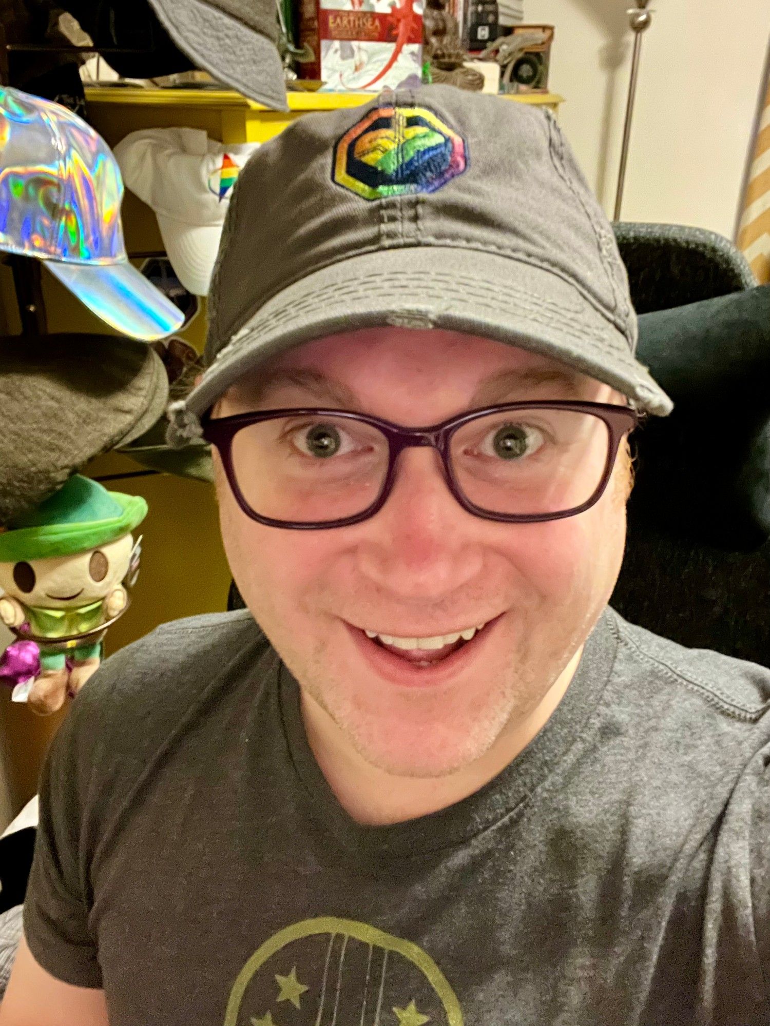 Selfie of a white guy looking excited. He is wearing dark glasses, a grey t-shirt, and a worn grey hat with a rainbow Streampunks logo. Behind him is a hat rack with various hats and a plushie on it and a shelf with a copy of Earthsea on it.