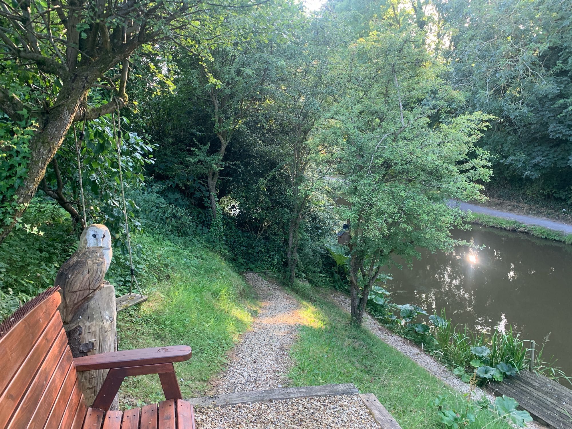 Garden path in dappled sunlight next a bench and a wooden statue of an owl, leading down to a canal which has sunlight reflecting off it