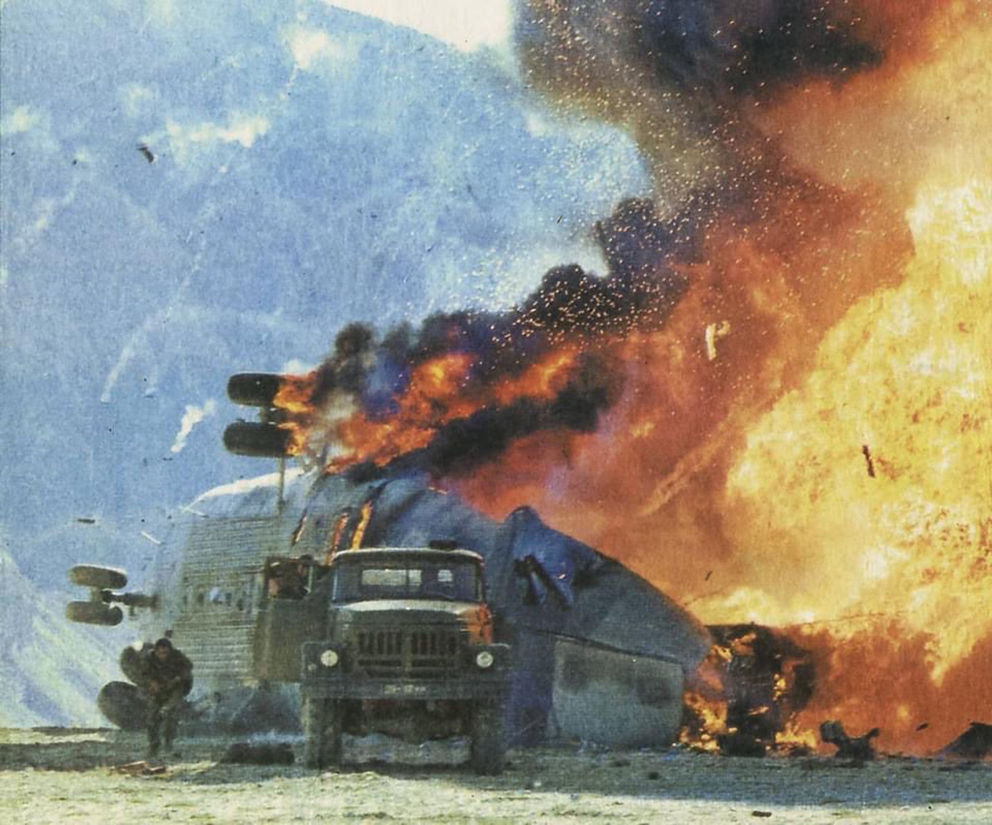 a somewhat lo-res ground-level photo of the rear of a very large helicopter, tipped over on its starboard side. there is a very strong fire engulfing it, from its port undercarriage at the highest point, through and across to its tail section, taking up about a third of the photo. debris in the air seems to imply a very recent explosion had occurred. In the foreground, there is a military truck facing head-on to the camera, with the passenger door open, and a person running away from the copter alongside the truck. are they possible carrying someone on their back(?)