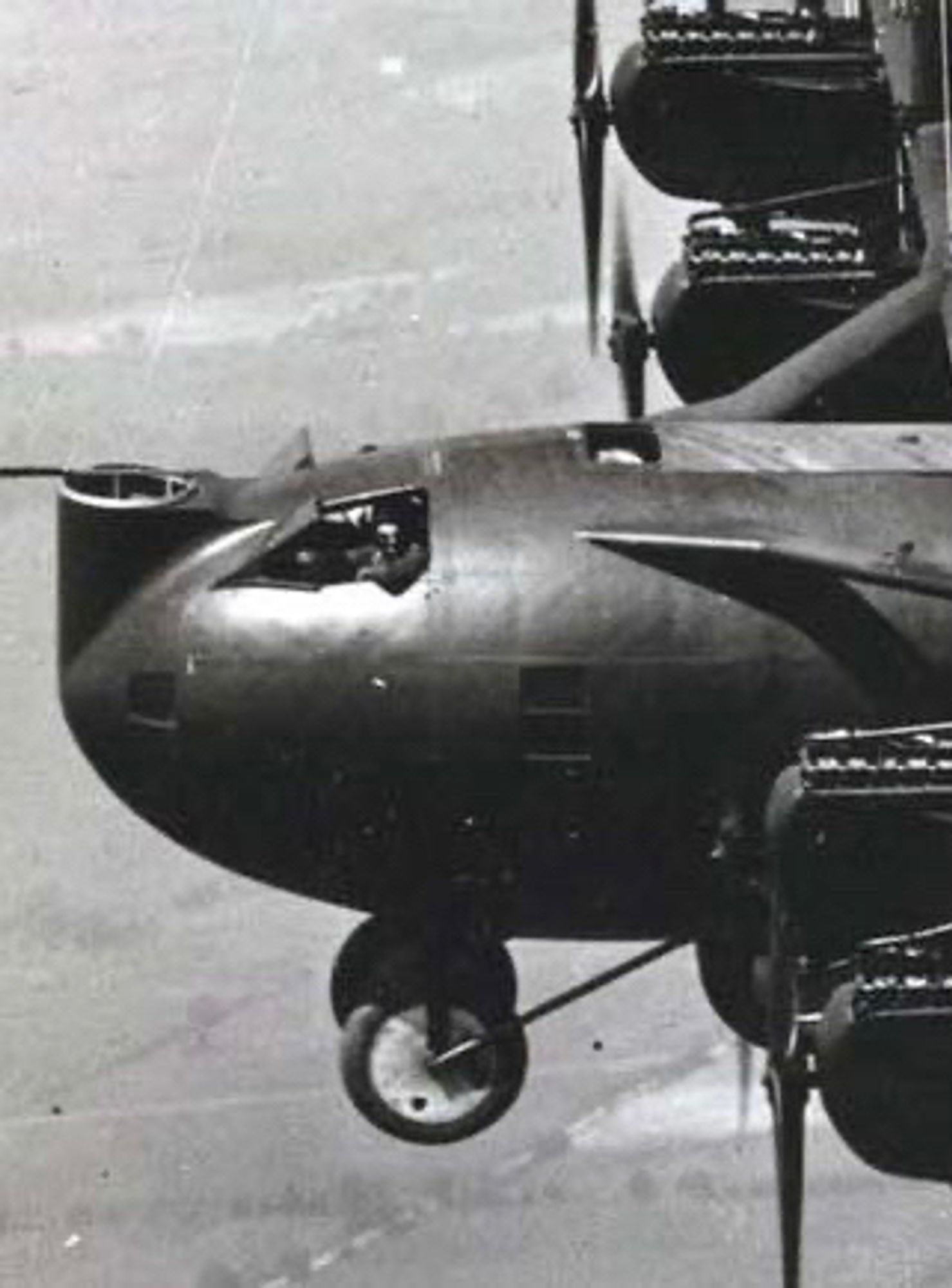 black and white photo of the front of the giant Witteman-Lewis XNBL-1 bomber in flight. a crew member - the pilot? is casually leaning out of the port cockpit, his elbow hanging over the side. in front of him, we have an empty gun turret, and the large fixed nose undercarriage is framed against fields far below